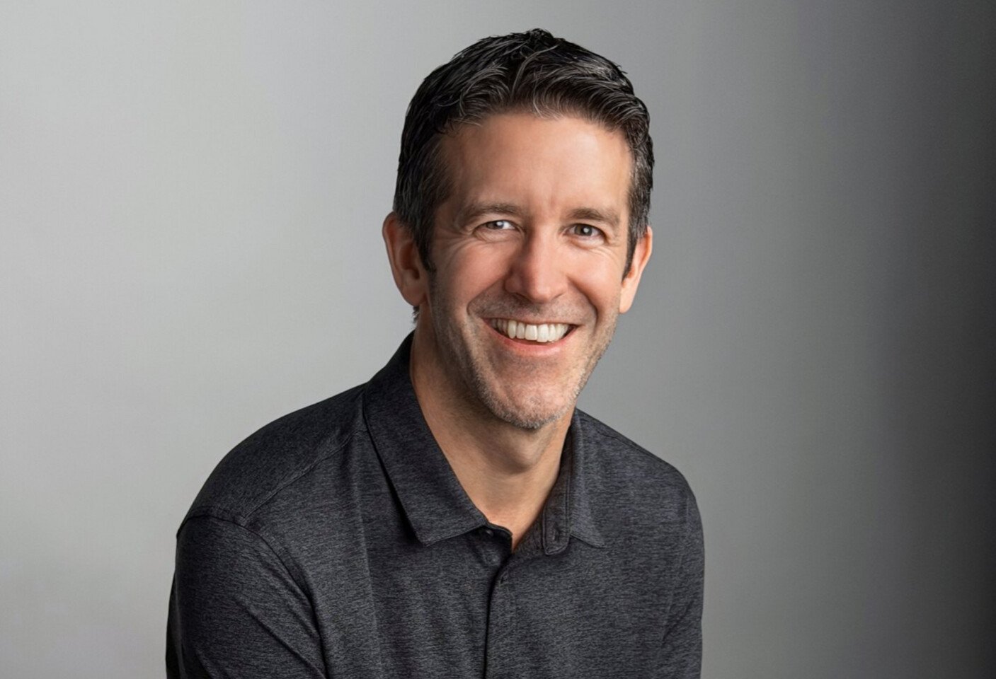Smiling man with short dark hair in a charcoal polo photographed against a gray studio backdrop