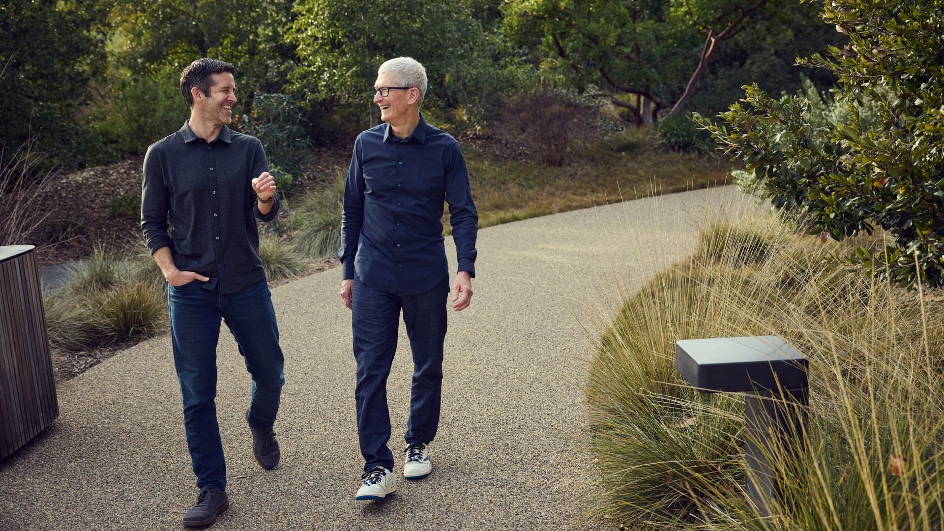 Two men walk side by side along a gravel path in a park smiling and talking as they stroll