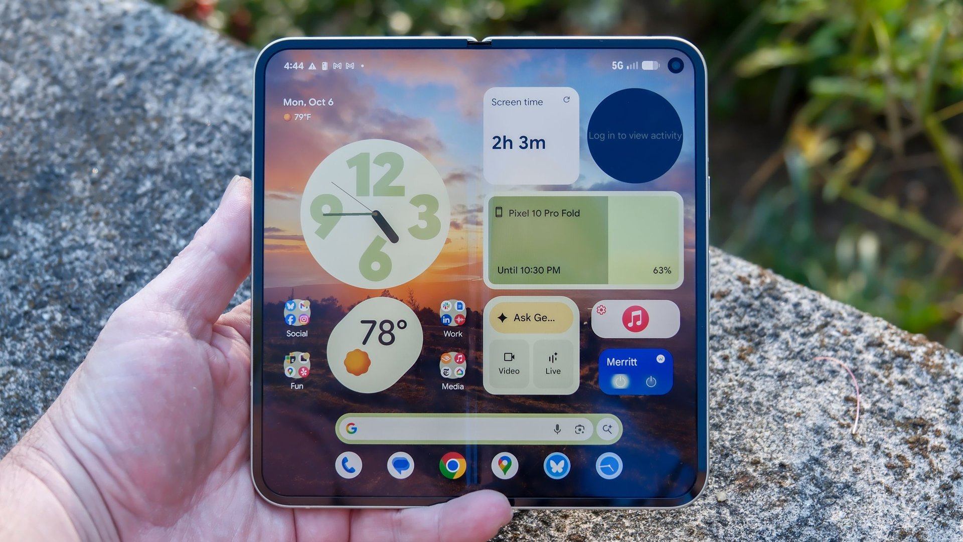 Close up of a hand holding a foldable smartphone outdoors on a rock showing a colorful home screen with widgets and a sunset wallpaper