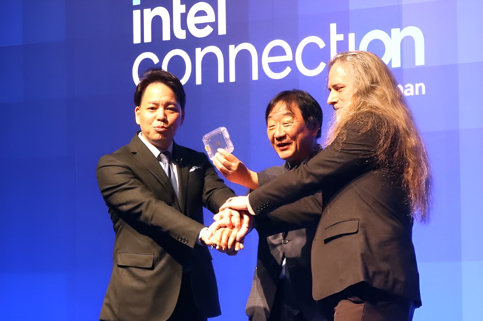 Three men in suits join hands in a group handshake on a blue stage with intel connection backdrop