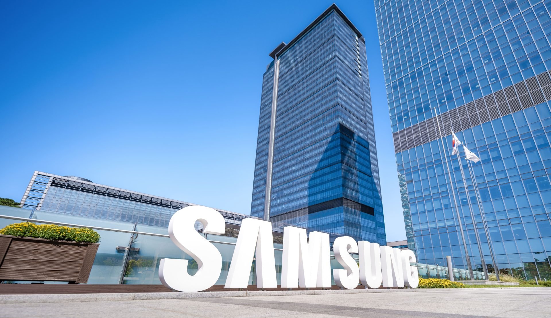 Exterior view of Samsung headquarters with large white SAMSUNG letters in the foreground and blue glass skyscrapers against a clear sky