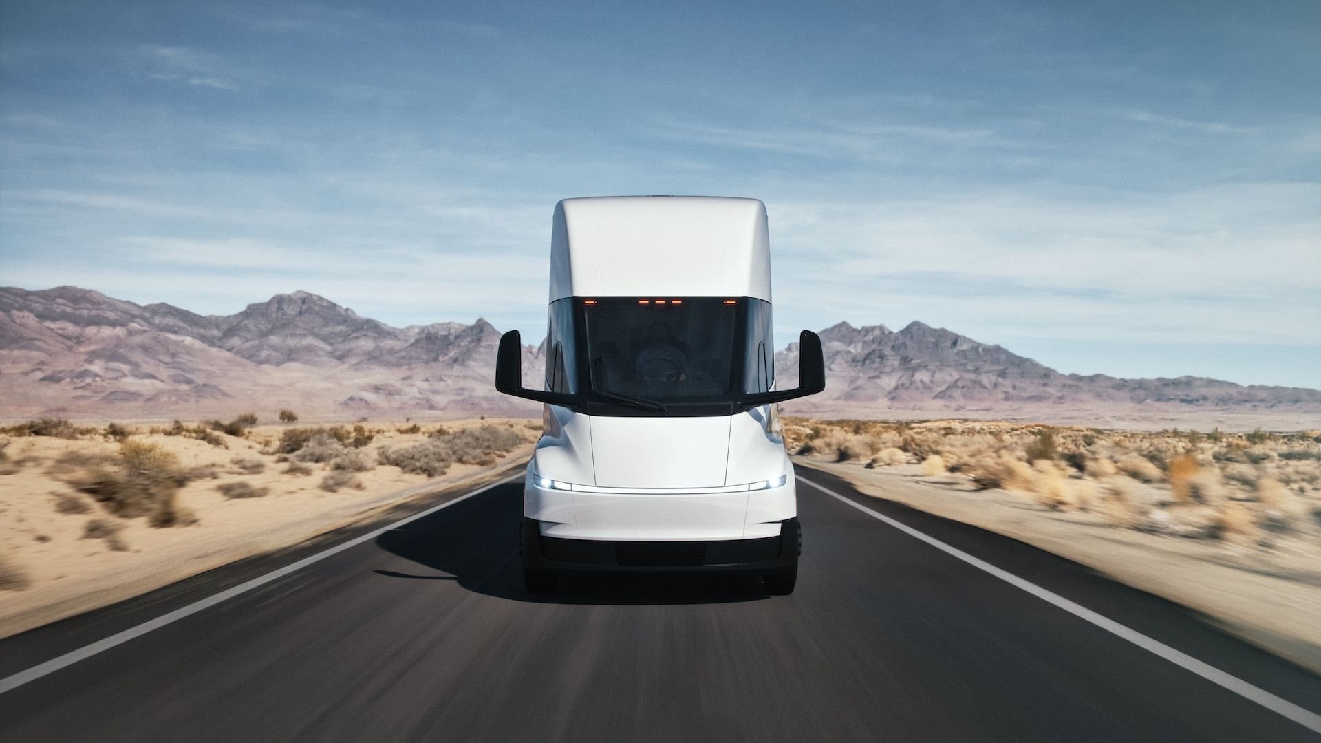 Front view of a large white semi truck driving toward the camera on a desert highway with mountains in the distance