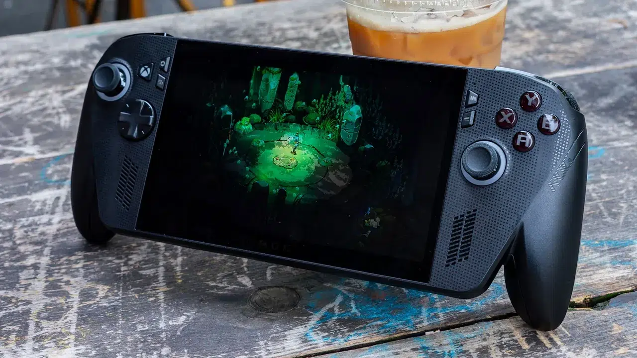 Handheld game console on a weathered wooden table showing a green lit fantasy arena on its screen with a glass of beer in the background