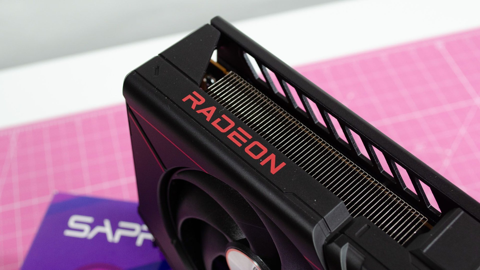 Close up of a black AMD Radeon graphics card with the red RADEON logo and exposed cooling fins on a pink background mat