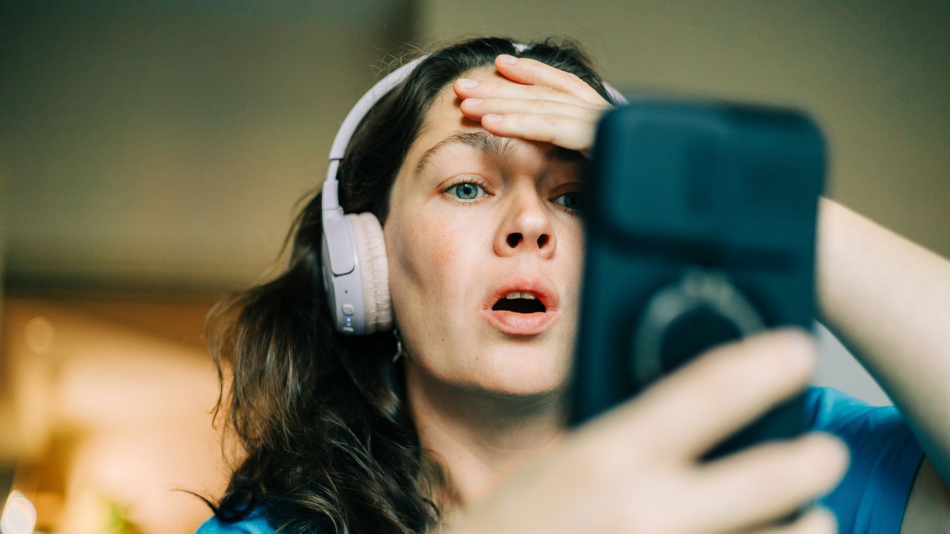 Woman wearing white over ear headphones looks surprised while taking a selfie with a smartphone held in front of her face