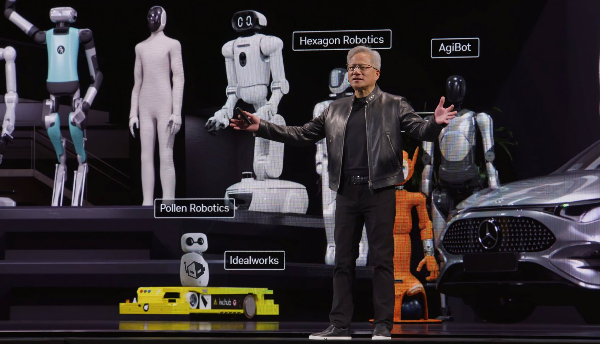 Man in a leather jacket presenting on a stage with multiple humanoid robots and a Mercedes car in the background labeled with company names like Hexagon Robotics and Pollen Robotics