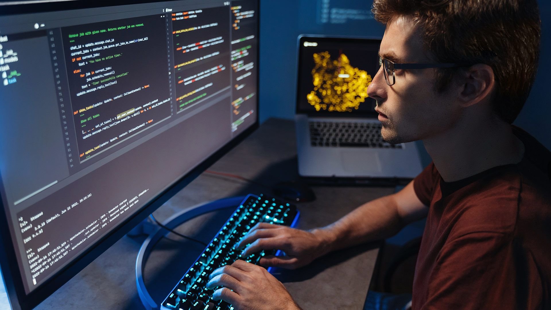 Programmer typing at a multi screen setup with code on the left monitor and a laptop in the background blue keyboard glow