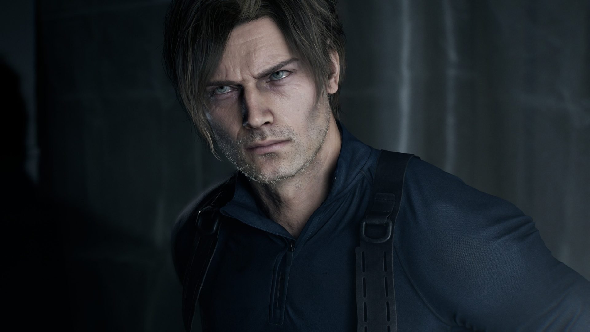Close up of a man with blue eyes and stubble wearing a dark tactical jacket with straps in a dim setting