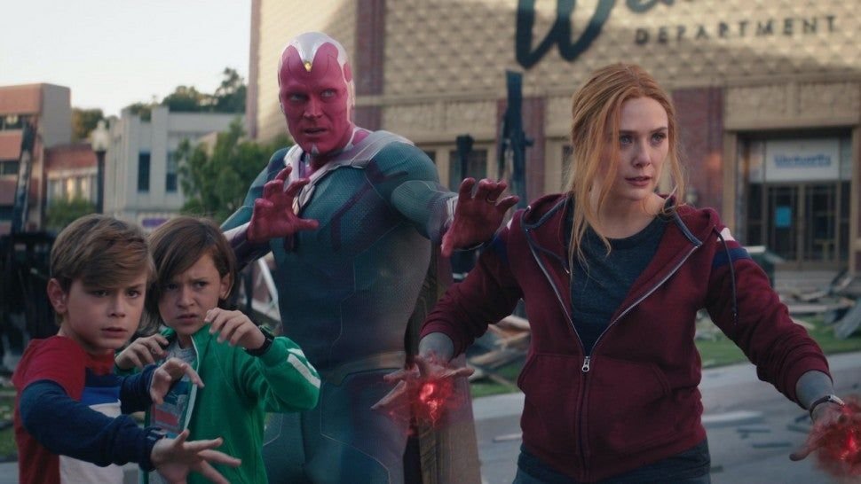 A teenage girl with glowing red energy in her hands stands at a street with two young boys a CGI superhero looming behind them guarding them