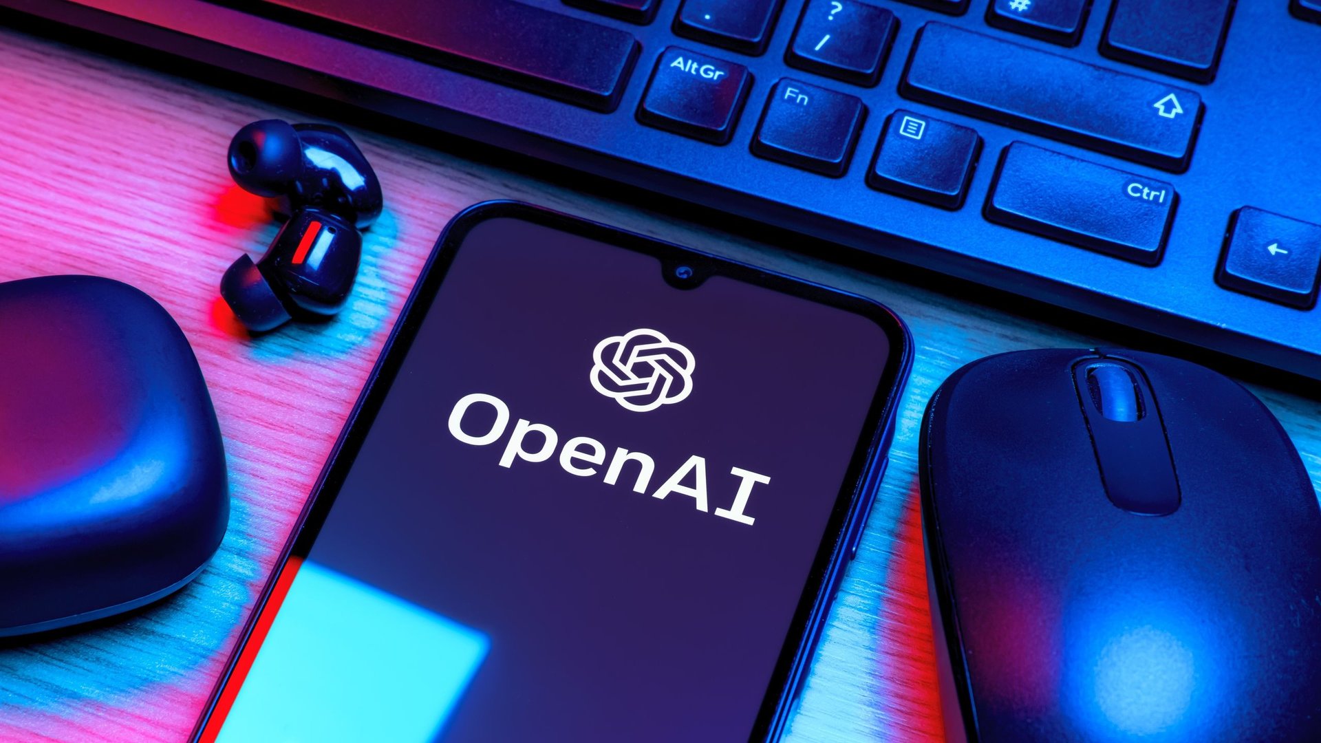 Smartphone showing the OpenAI logo on a desk with a keyboard mouse and wireless earbuds under colorful blue and pink lighting