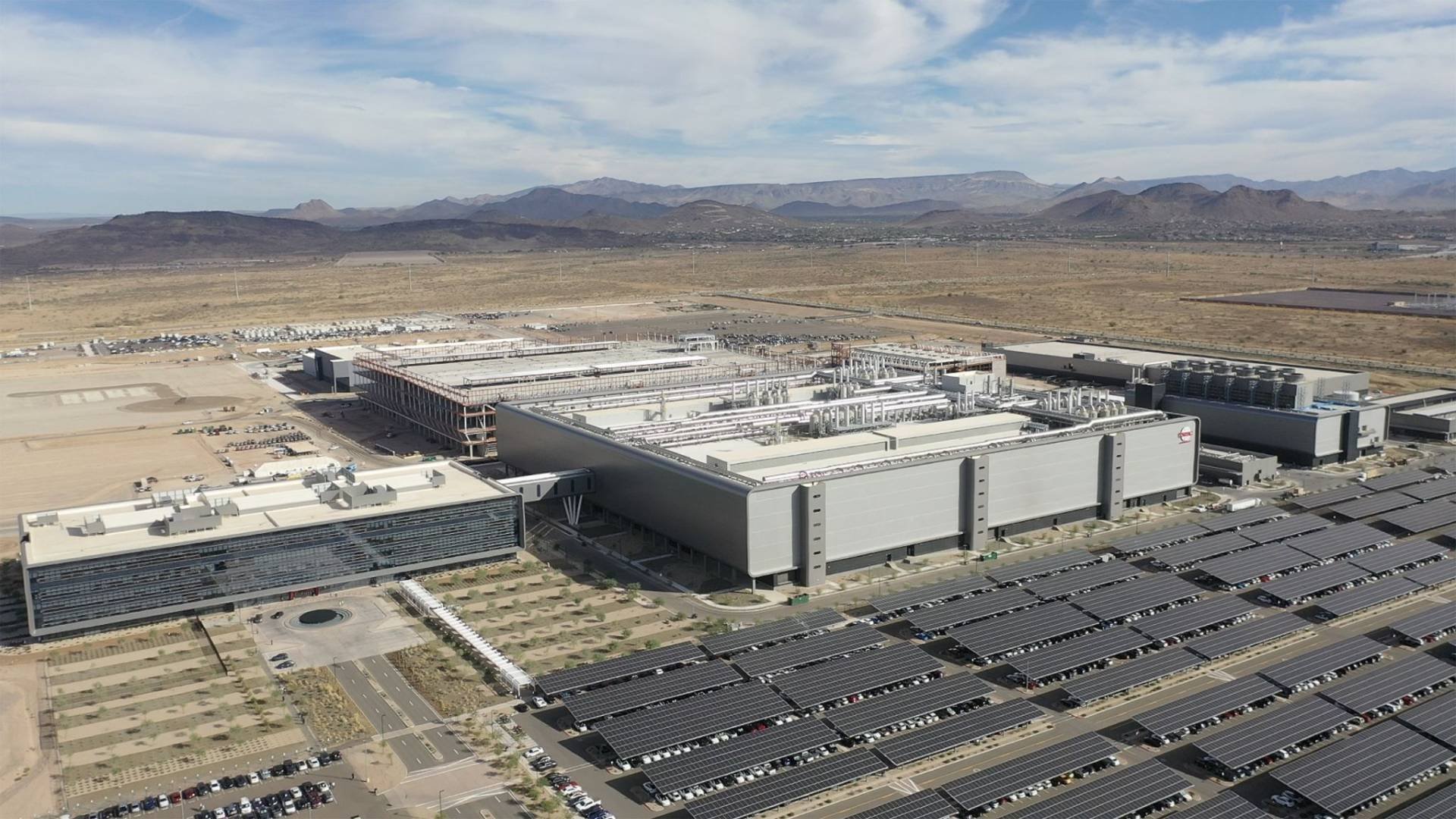 Industrial facility with extensive rooftop equipment and rows of solar panels in the parking lots desert landscape beyond