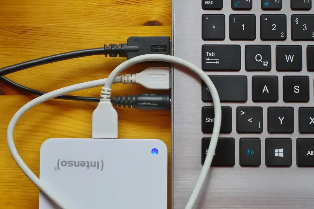 White external USB hard drive connected to a silver laptop with cables plugged in and a blue LED light on the drive