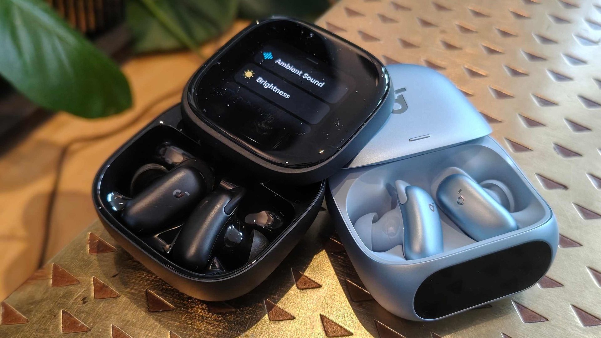 Two wireless earbud cases open a black case with black earbuds and a blue case with light blue earbuds on a metallic patterned surface