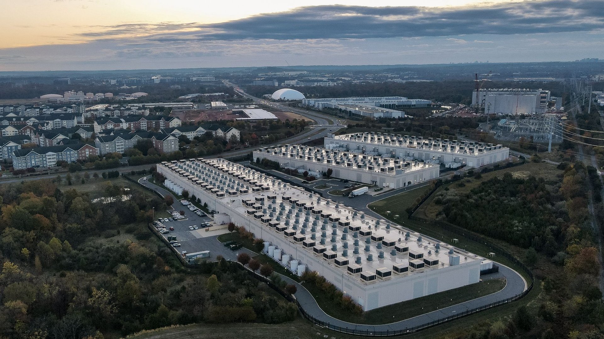 Aerial view of a large data center campus with multiple white buildings and rooftop cooling units surrounded by trees and a nearby cityscape at duskearly morning light