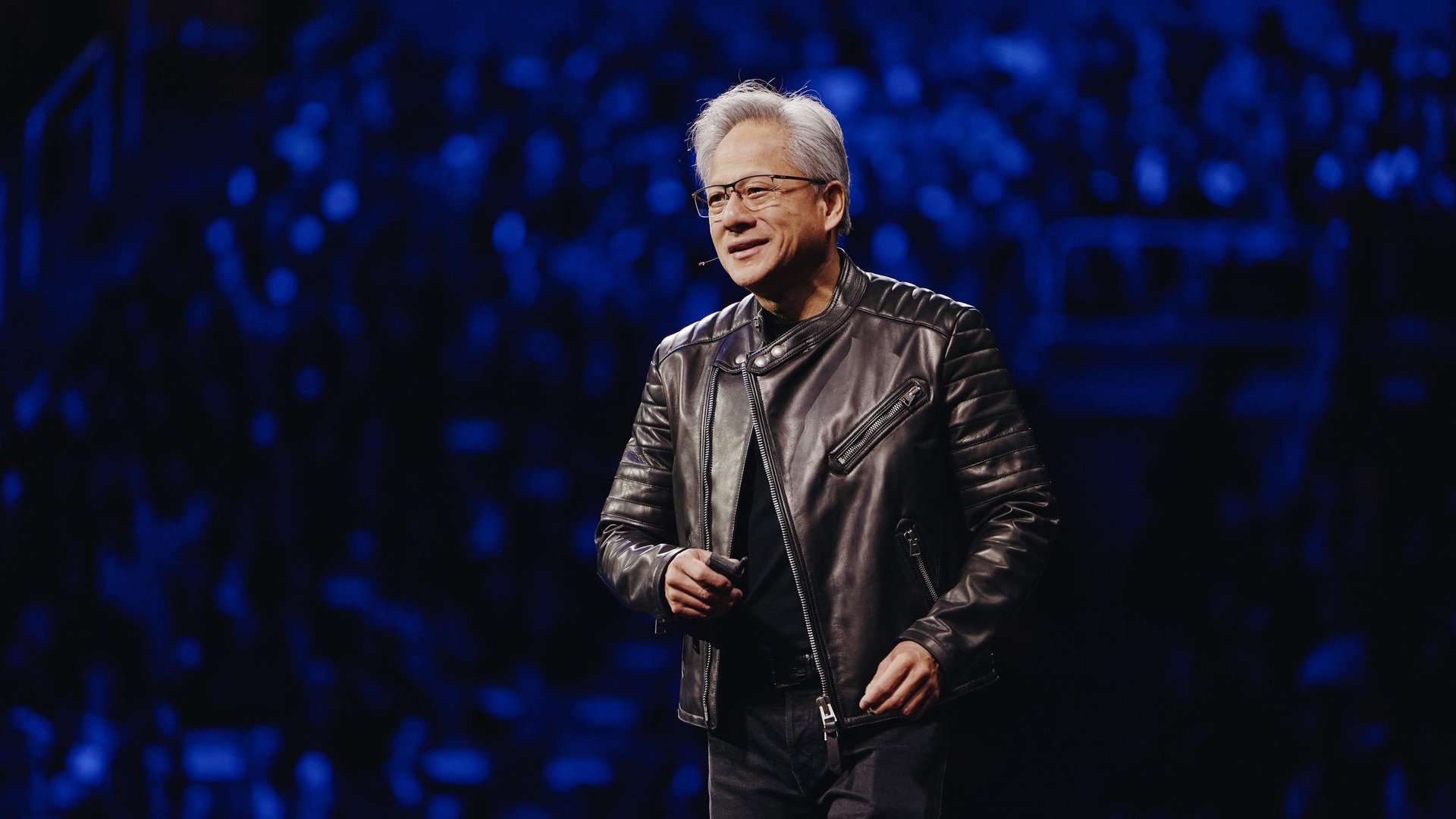 Speaker in a black leather jacket on stage addressing a blue lit audience