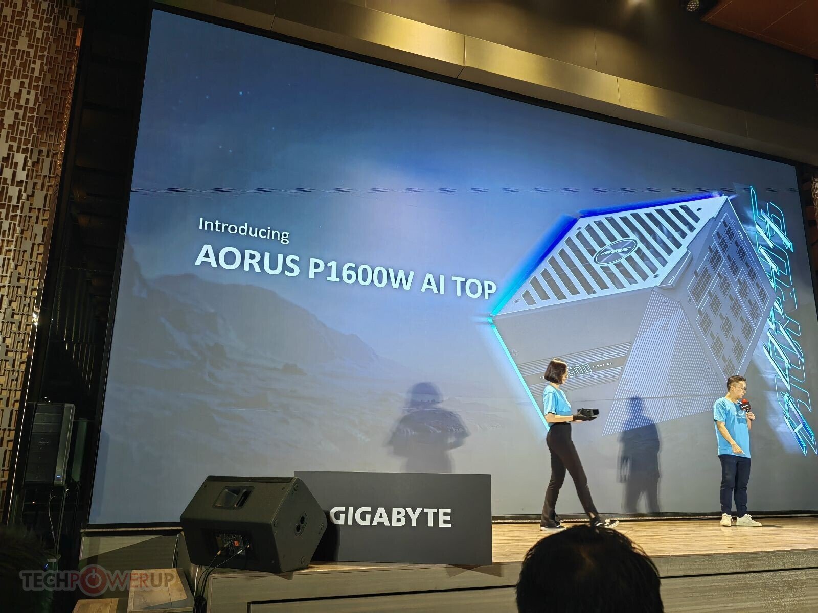 Stage presentation announcing AORUS P1600W AI TOP on a large screen two hosts in blue shirts speak on stage with Gigabyte branding visible