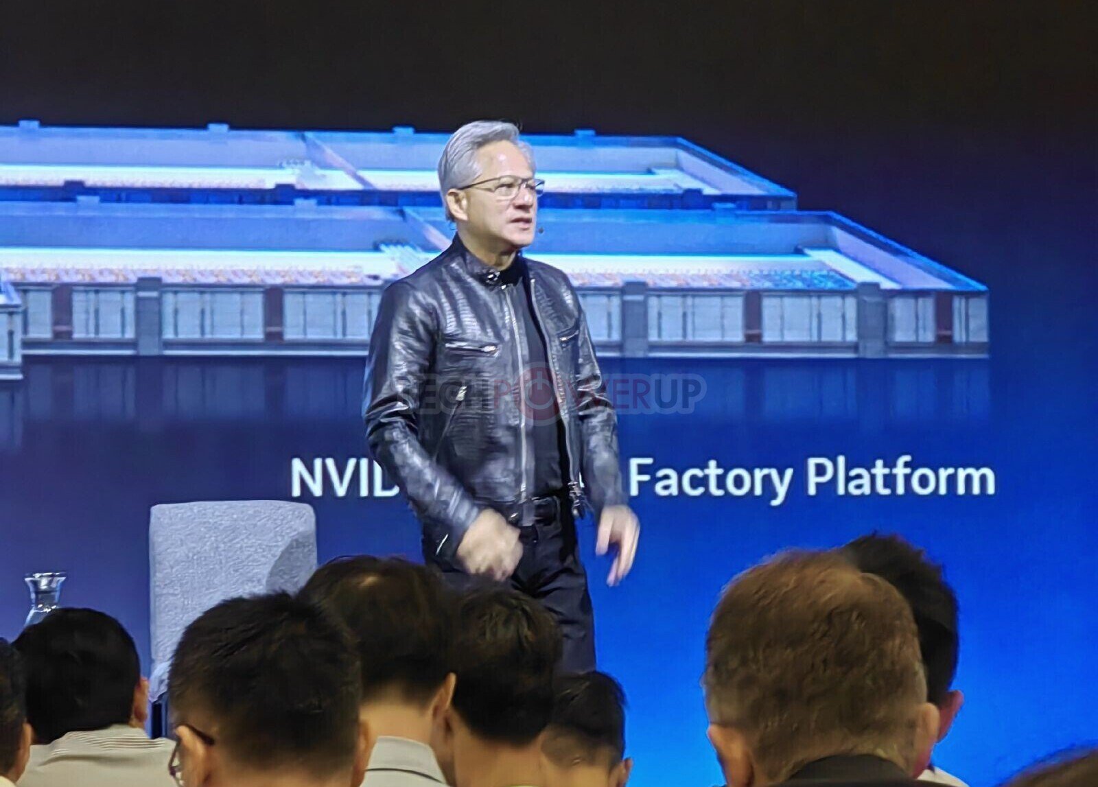 Speaker in a black leather jacket presenting on a stage with a blue backdrop reading NVIDIA Factory Platform