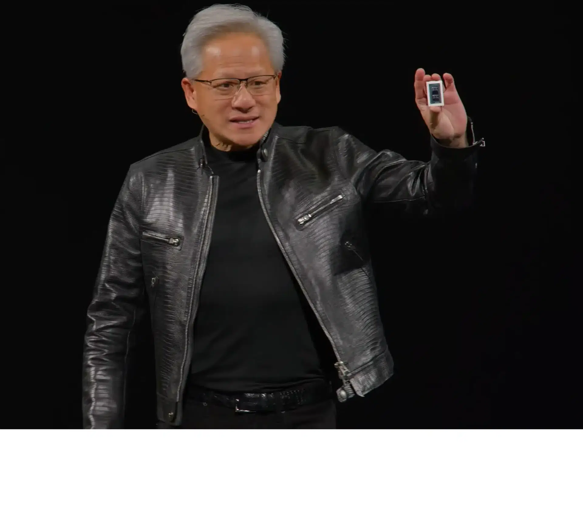 Tech keynote speaker with gray hair and glasses in a black leather jacket holding up a small handheld device against a dark background