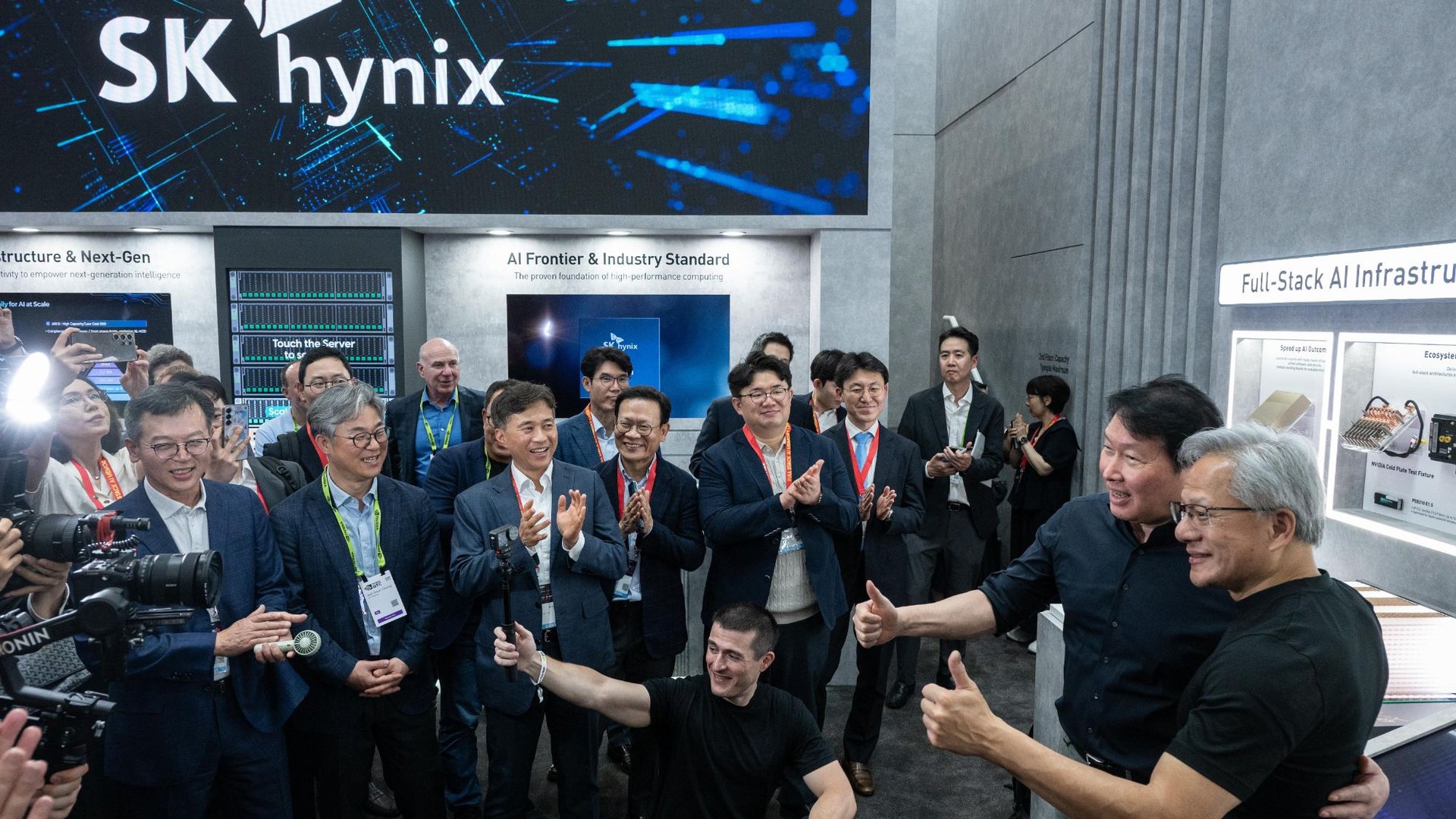 Group of professionals at a tech expo posing for photos with cameras and thumbs up in front of display panels