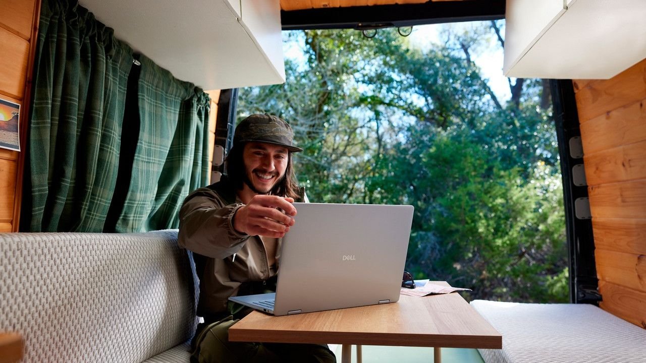 Man smiling and using a Dell laptop inside a wood paneled camper van with a small table and outdoor green scenery visible