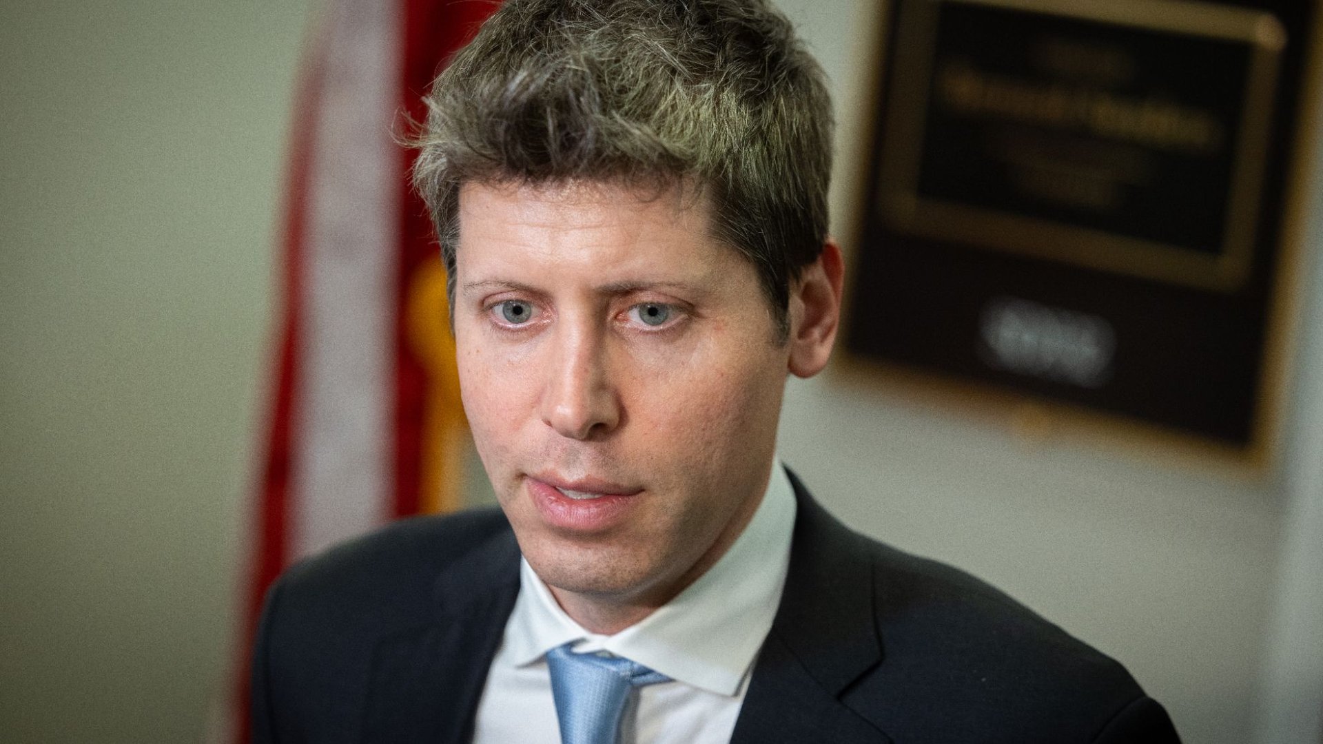 Portrait of a man in a dark suit with a light blue tie looking slightly downward in an indoor setting