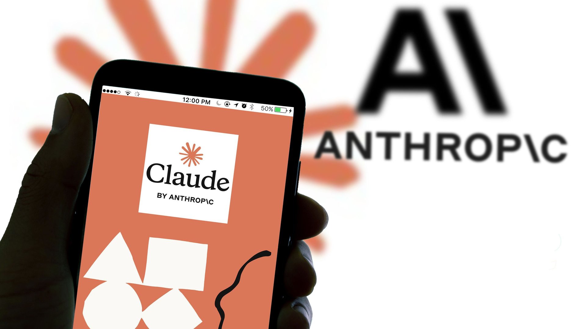 Hand holding a smartphone showing the Claude by Anthropic app logo on an orange background with a blurred Anthrop IC wordmark in the background