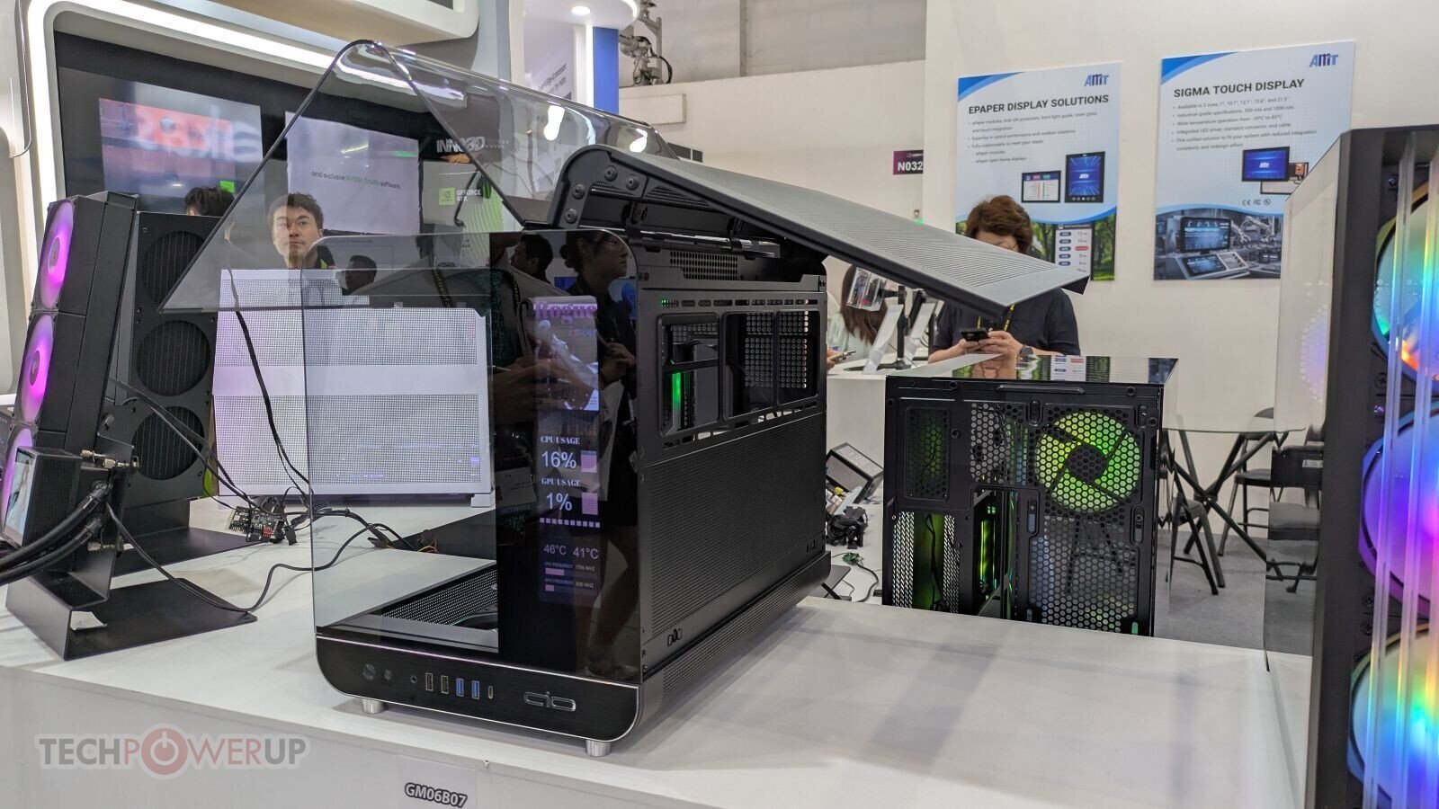 Open frame computer case with a large transparent side panel lifted open on a display table at a tech expo while people look on in the background