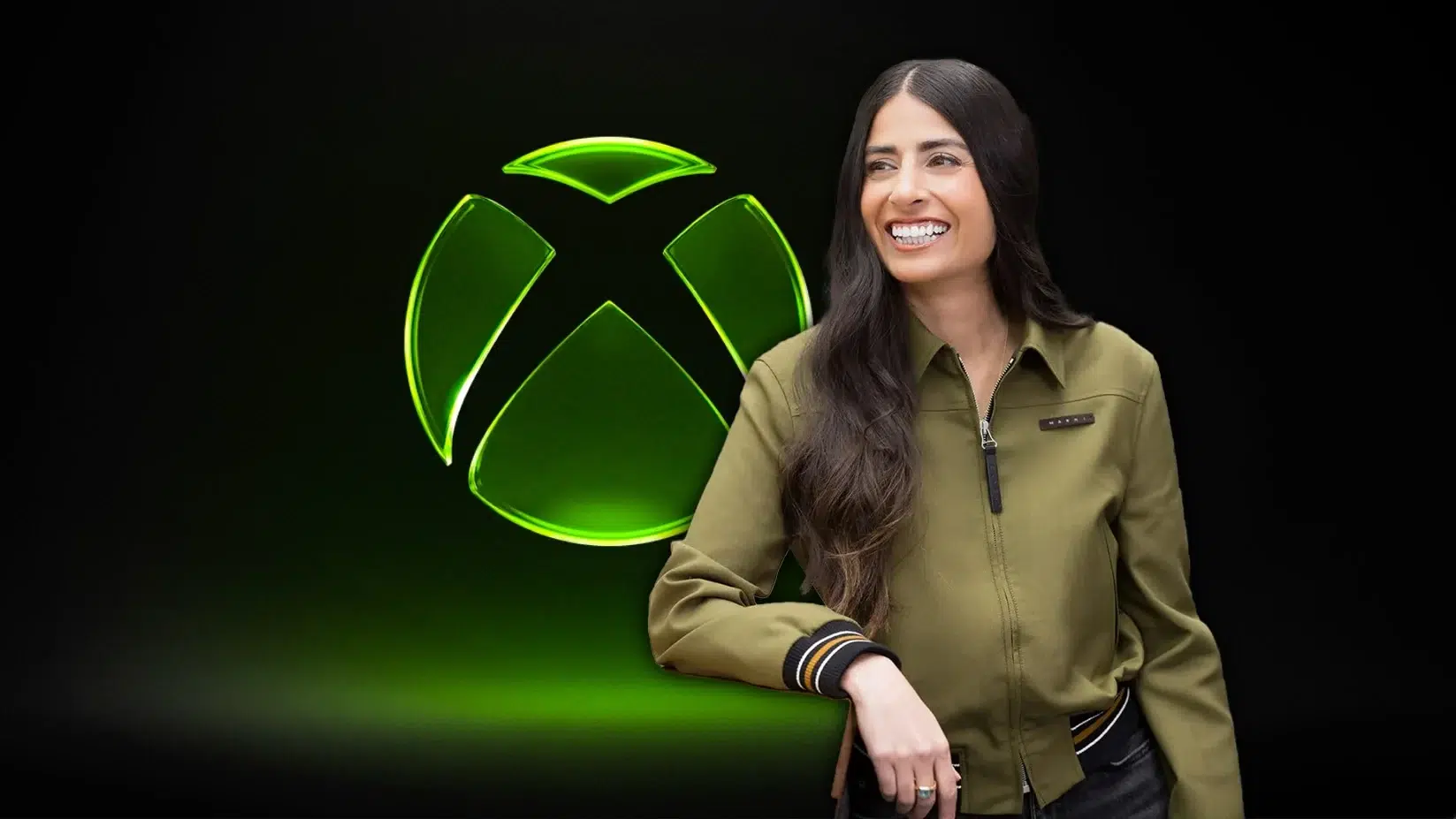 Smiling woman in olive jacket standing next to a large glowing green Xbox logo on a dark background