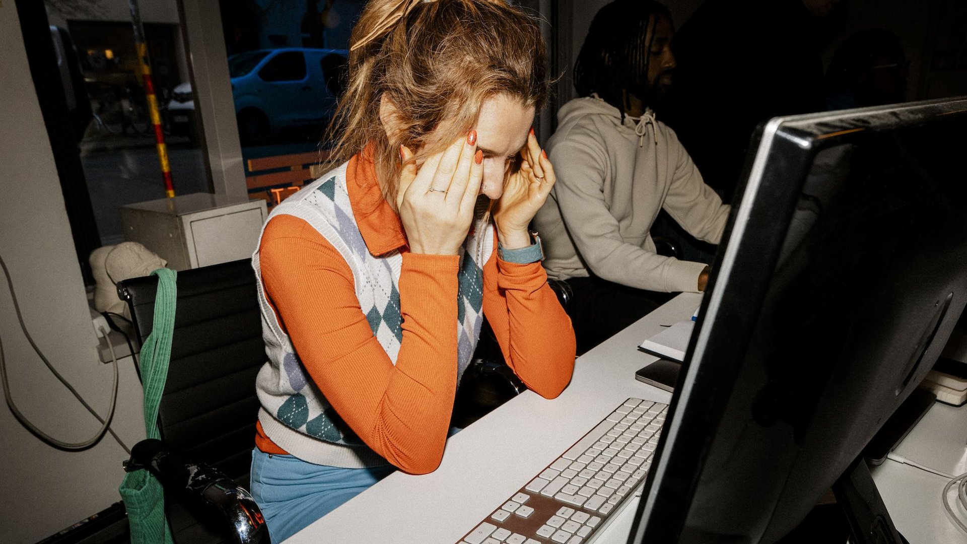 Woman at a desk in an office rubbing her temples in front of a computer screen showing stress or fatigue