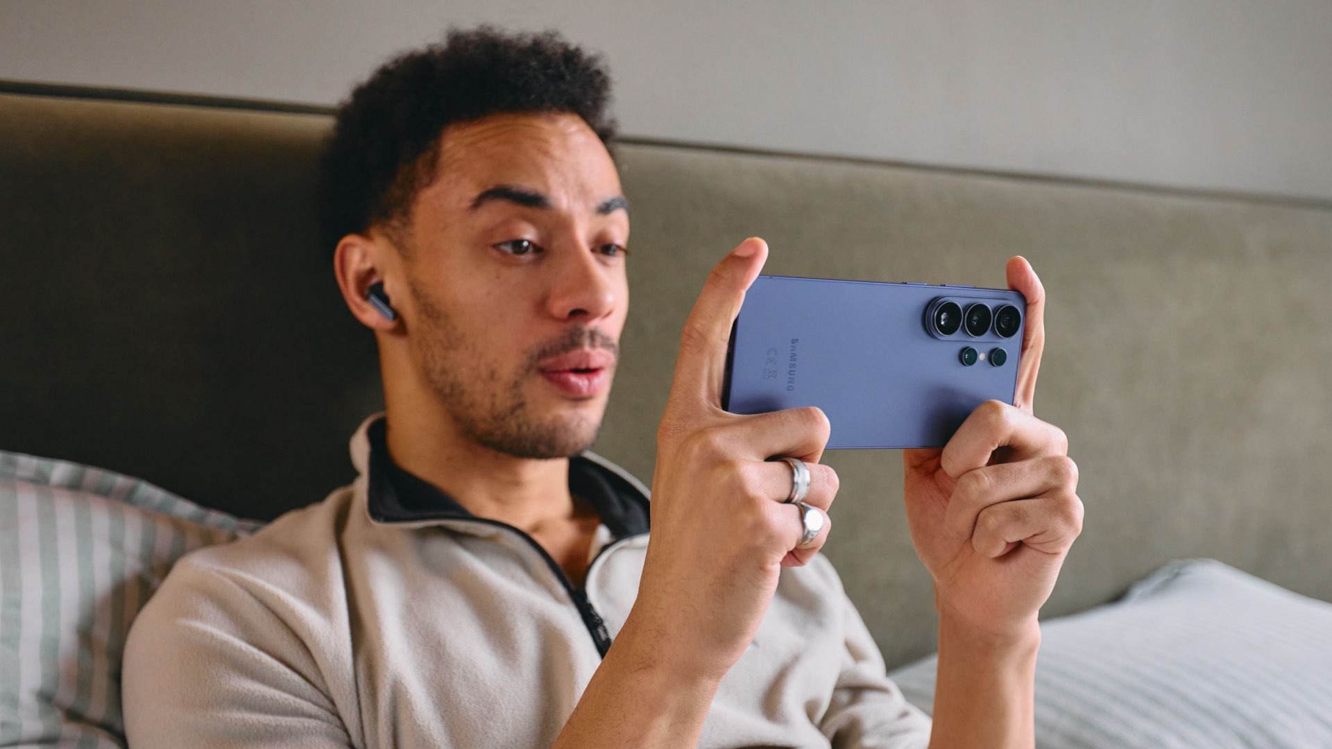Man lying in bed wearing a wireless earbud holding a blue Samsung phone with a multi camera array to take a photos selfie like shot