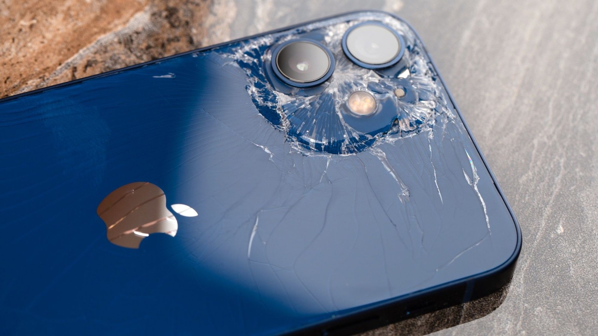 Blue iPhone with severely cracked back glass around the dual camera module after a drop showing shattered surface and scratches