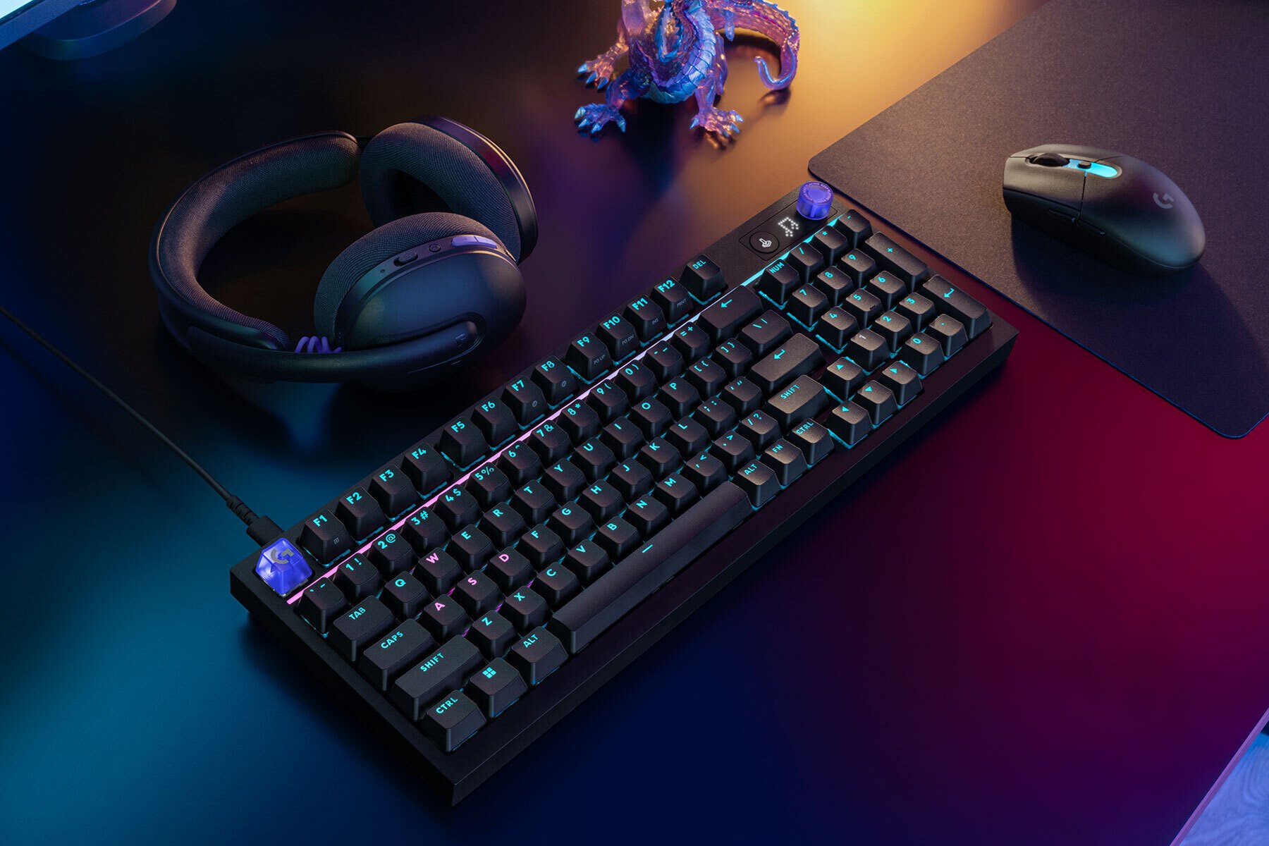 RGB mechanical keyboard with bluepurple backlighting on a dark desk alongside headphones a mouse and a small dragon figurine
