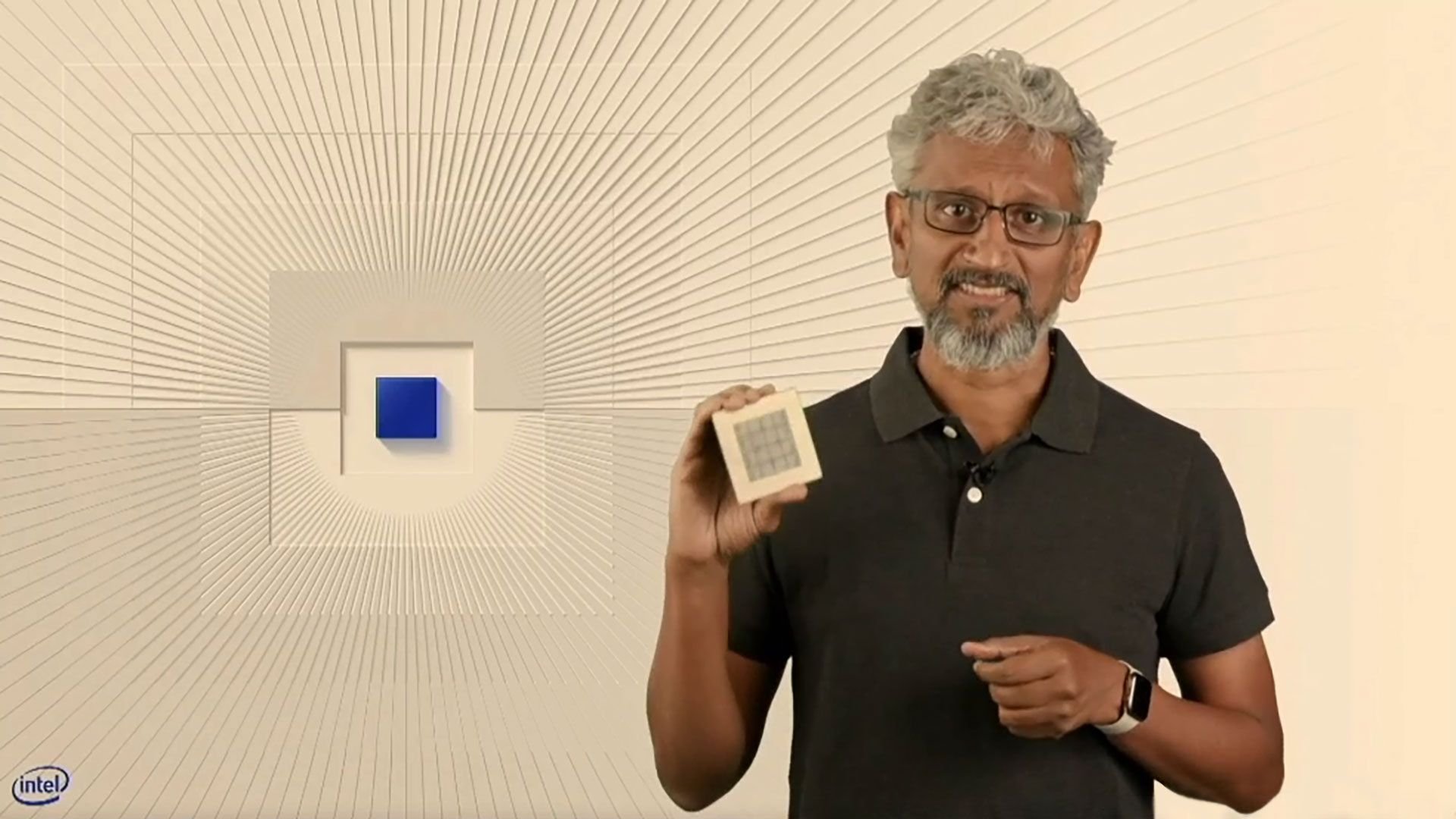 Man with gray hair and glasses holding up a small grid patterned tile in a bright geometric studio Intel logo visible in the corner