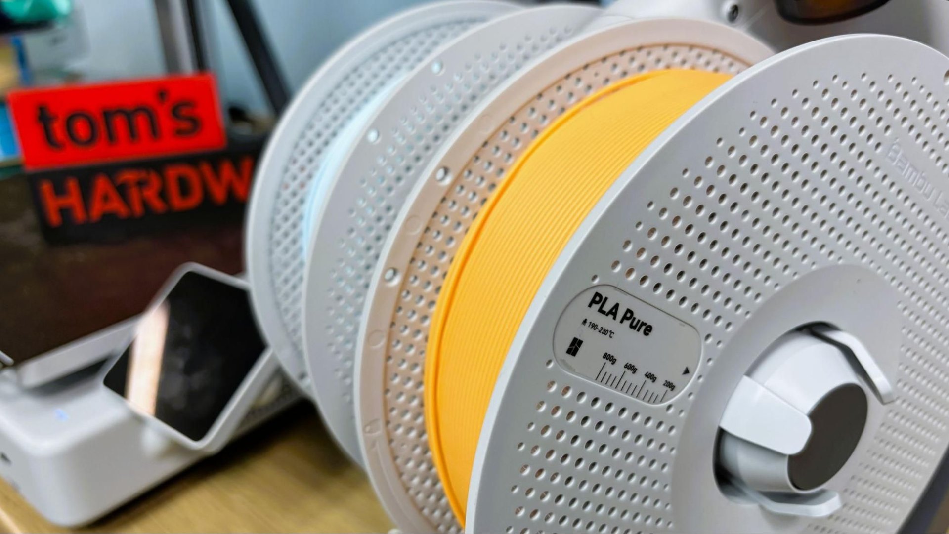 Close up of stacked 3D printer filament spools yellow PLA on a holder with a hardware store sign blurred in the background