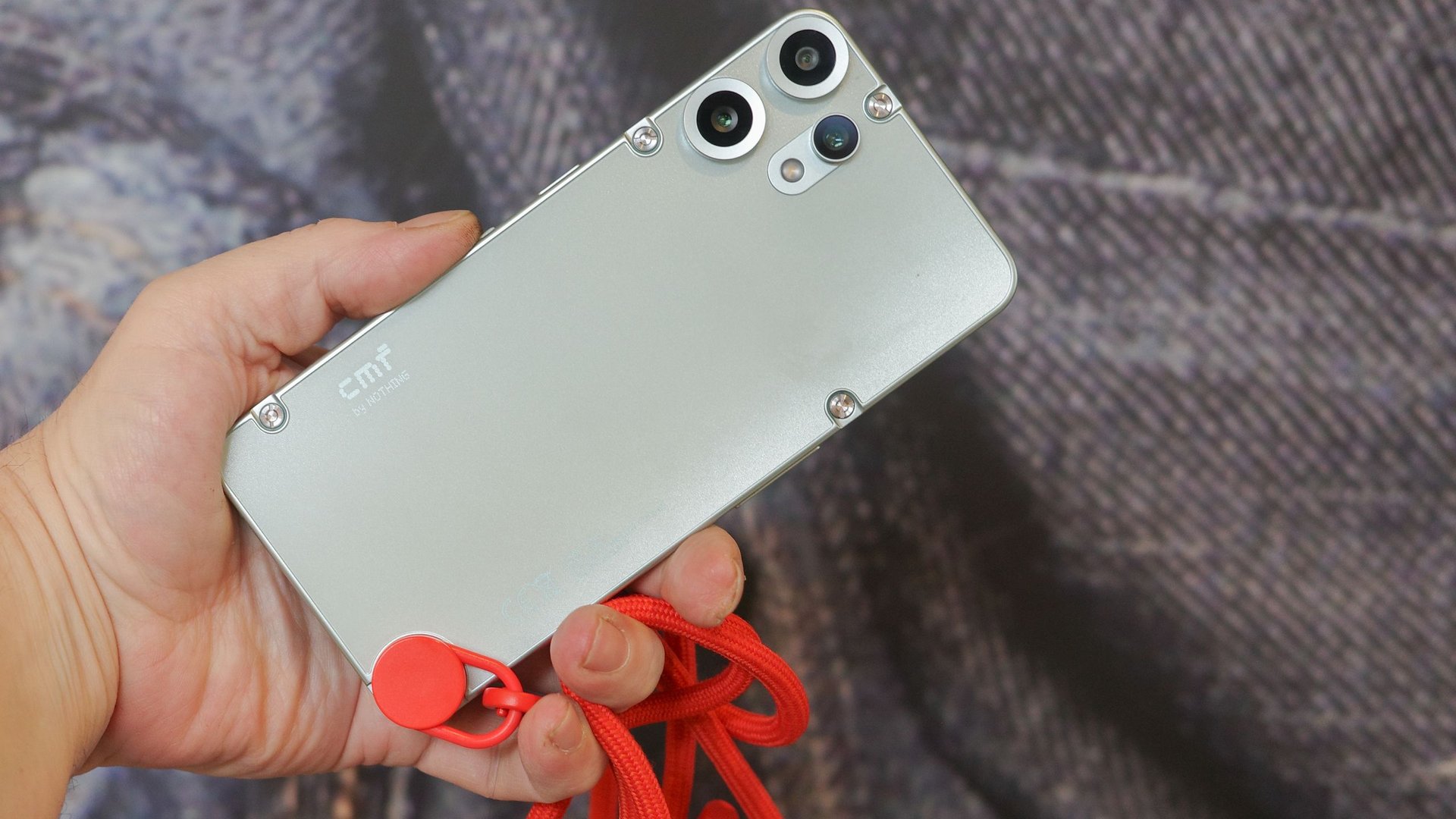 Person holding a silver smartphone with multiple rear cameras and a red lanyard attached to the bottom
