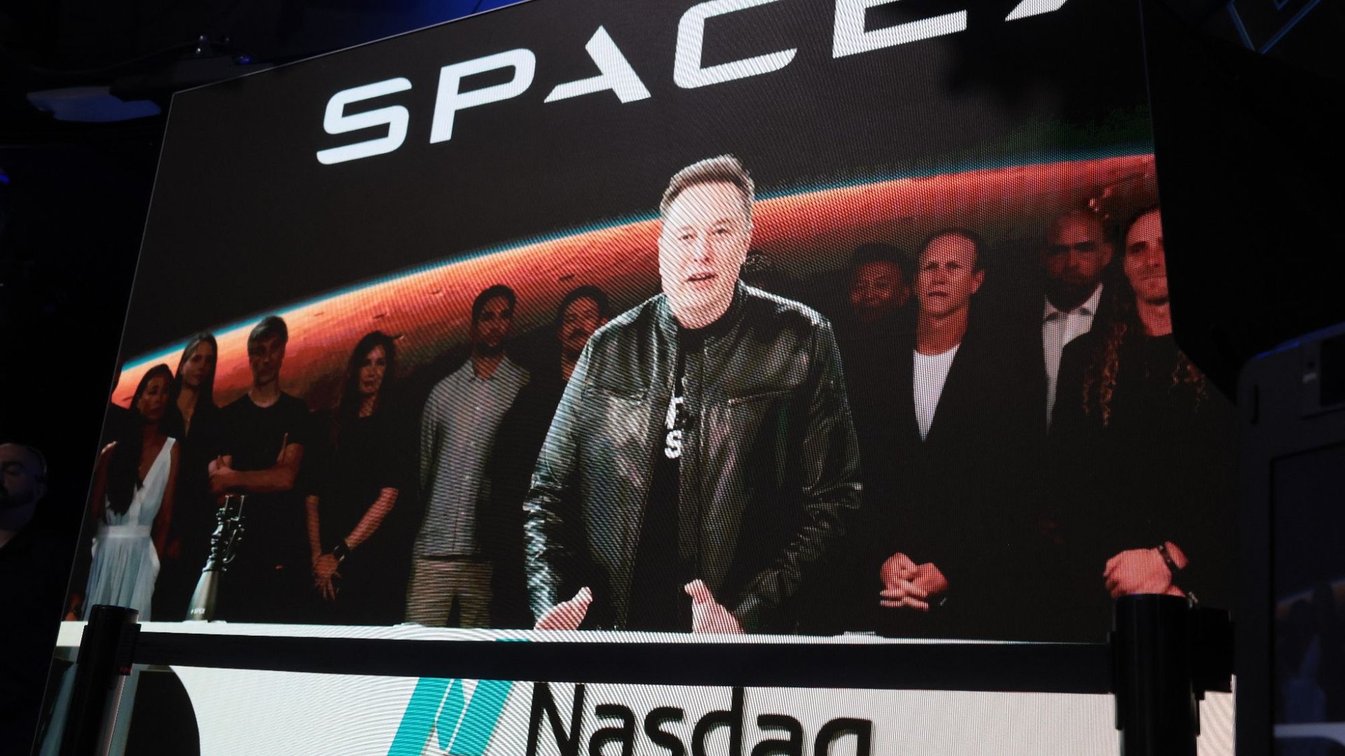 Large screen at a Nasdaq event showing a man in a leather jacket and a group of people behind him