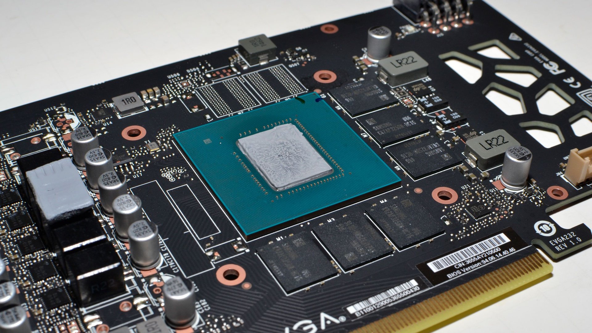 Close up of a graphics card PCB with a central GPU die mounted on a blue green chip surrounded by capacitors and memory chips