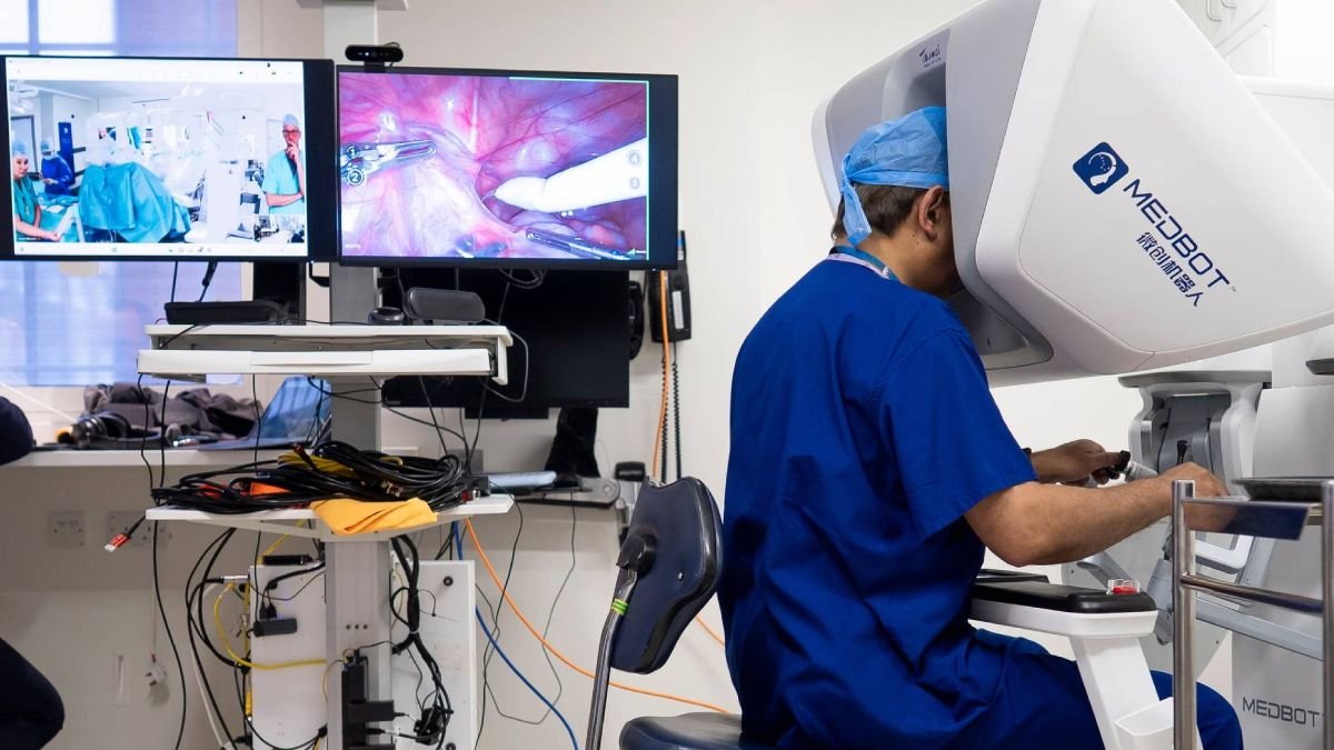 Surgeon in blue scrubs operates a robotic assisted surgery system with two monitors showing the procedure