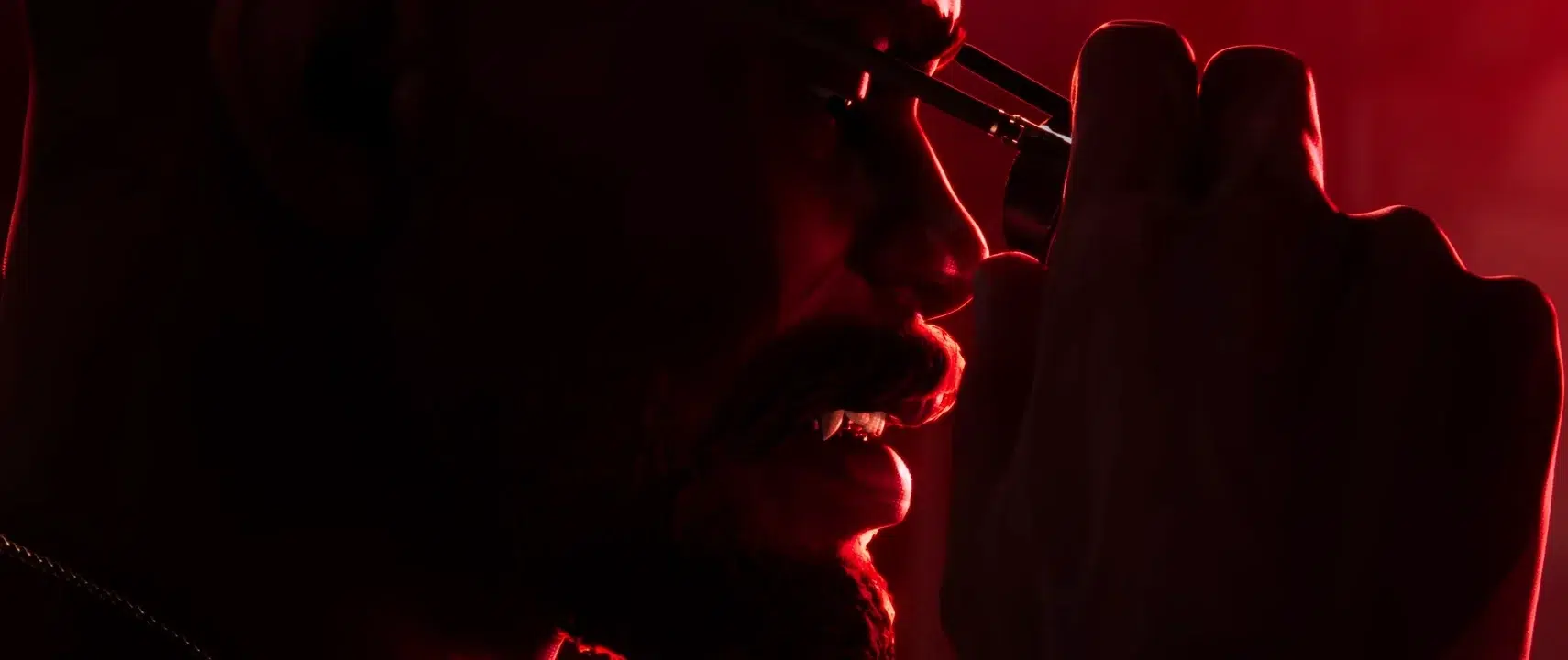 Close up of a hooded figure holding a metallic tool near their mouth bathed in red light