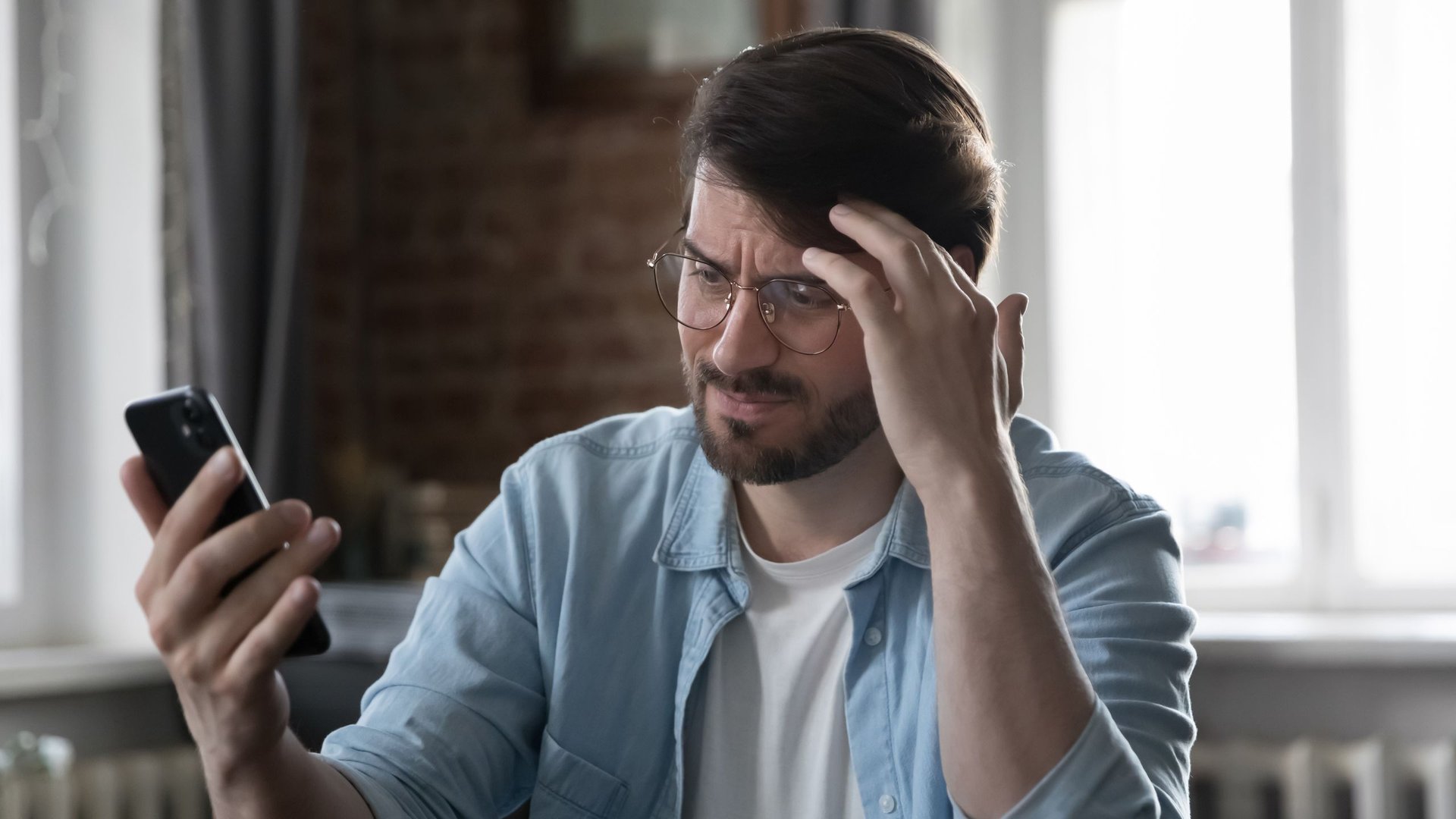 Man with glasses and a beard looks at his smartphone hand on temple puzzled