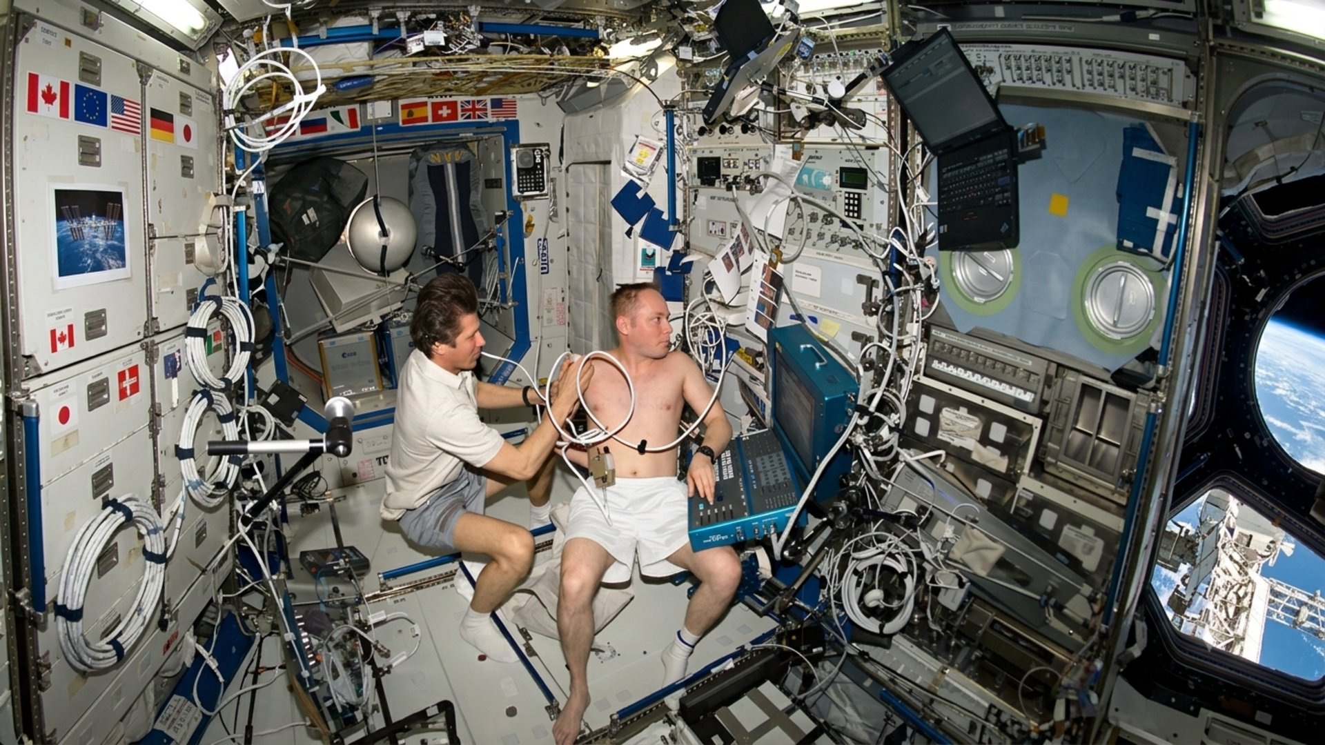 Two astronauts float inside a crowded ISS cabin surrounded by cables and flight panels with Earth visible outside the window
