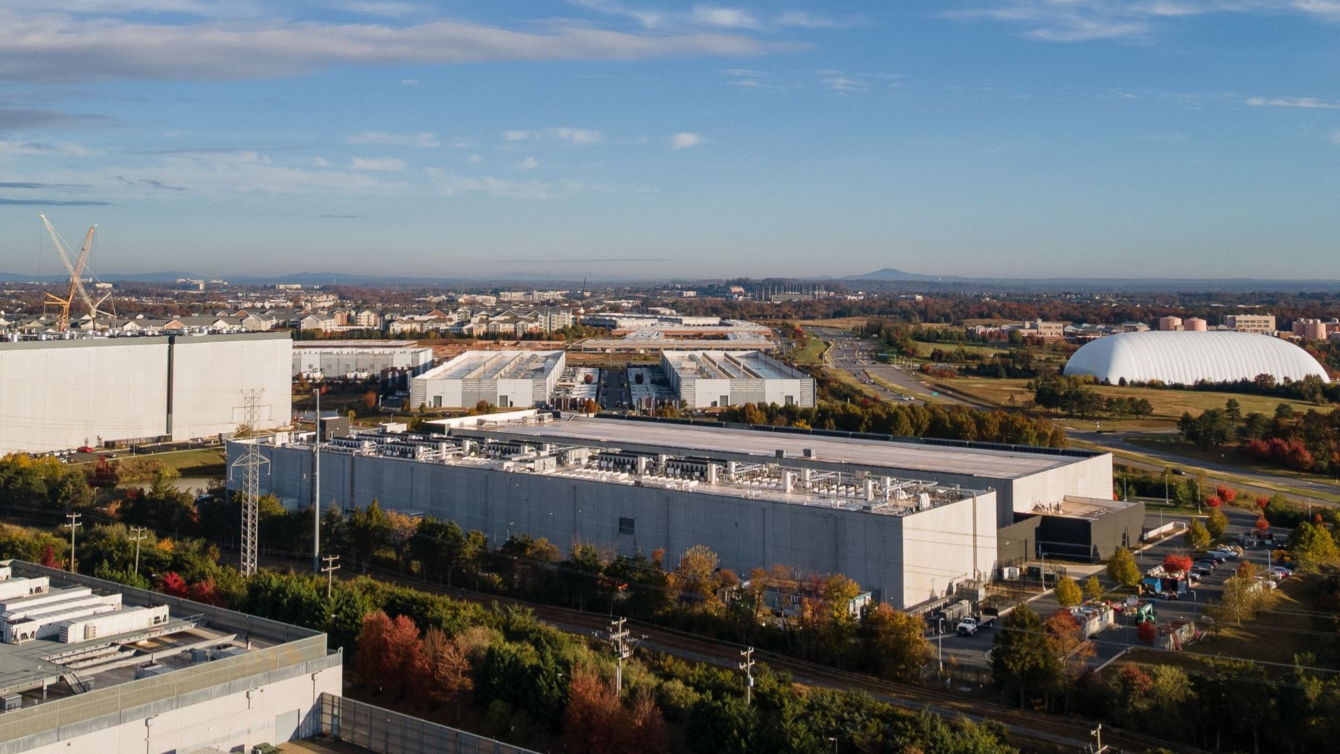 Aerial view of an industrial campus with large white warehouses a construction crane on the left and a large white dome shaped building on the right with autumn trees and a distant cityscape