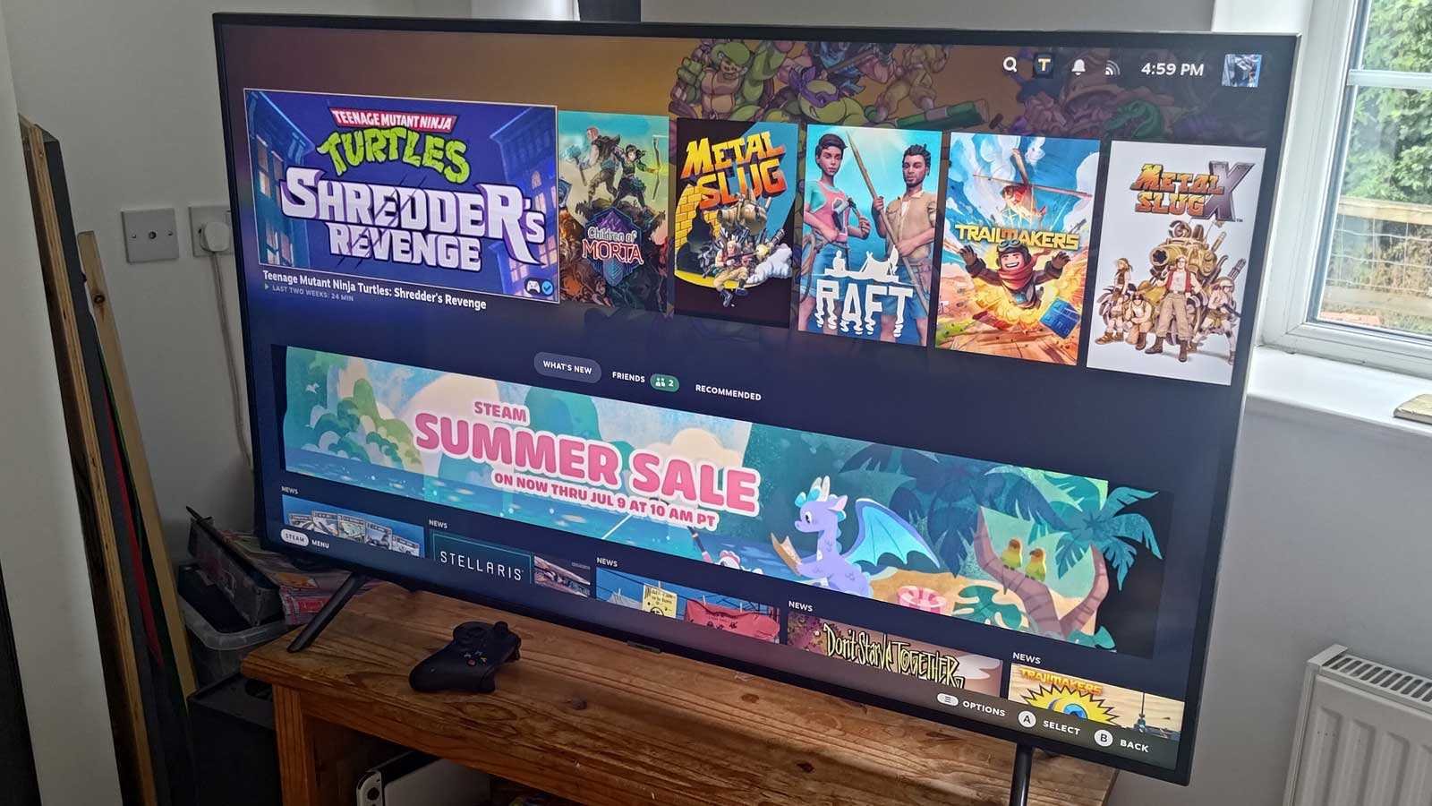 TV screen showing a Steam library with game tiles like Shredders Revenge and Metal Slug plus a colorful Summer Sale banner and a gamepad on a wooden table in a living room
