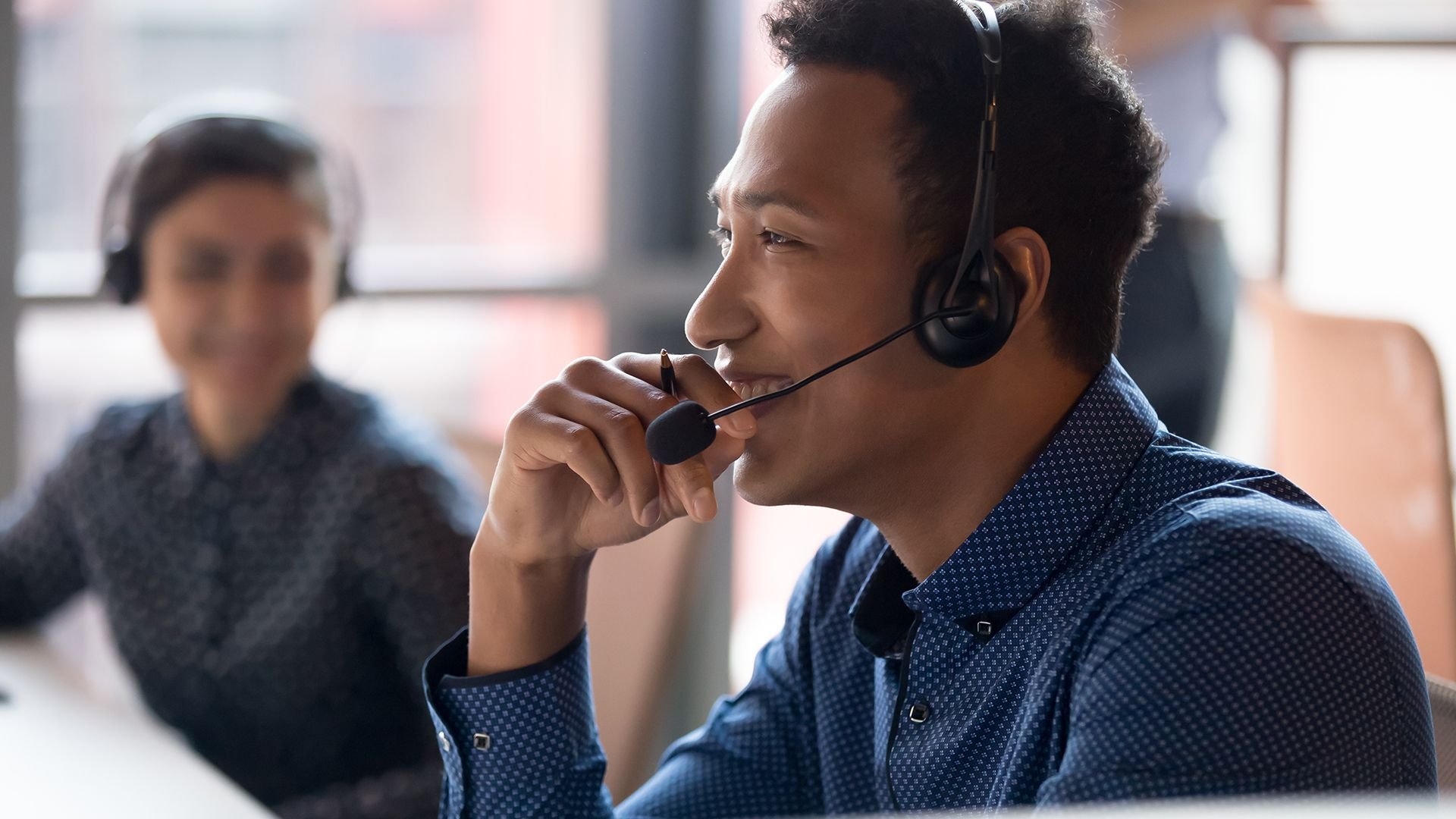 Man wearing a headset with microphone speaking at a desk in an office environment with a blurred colleague in the background