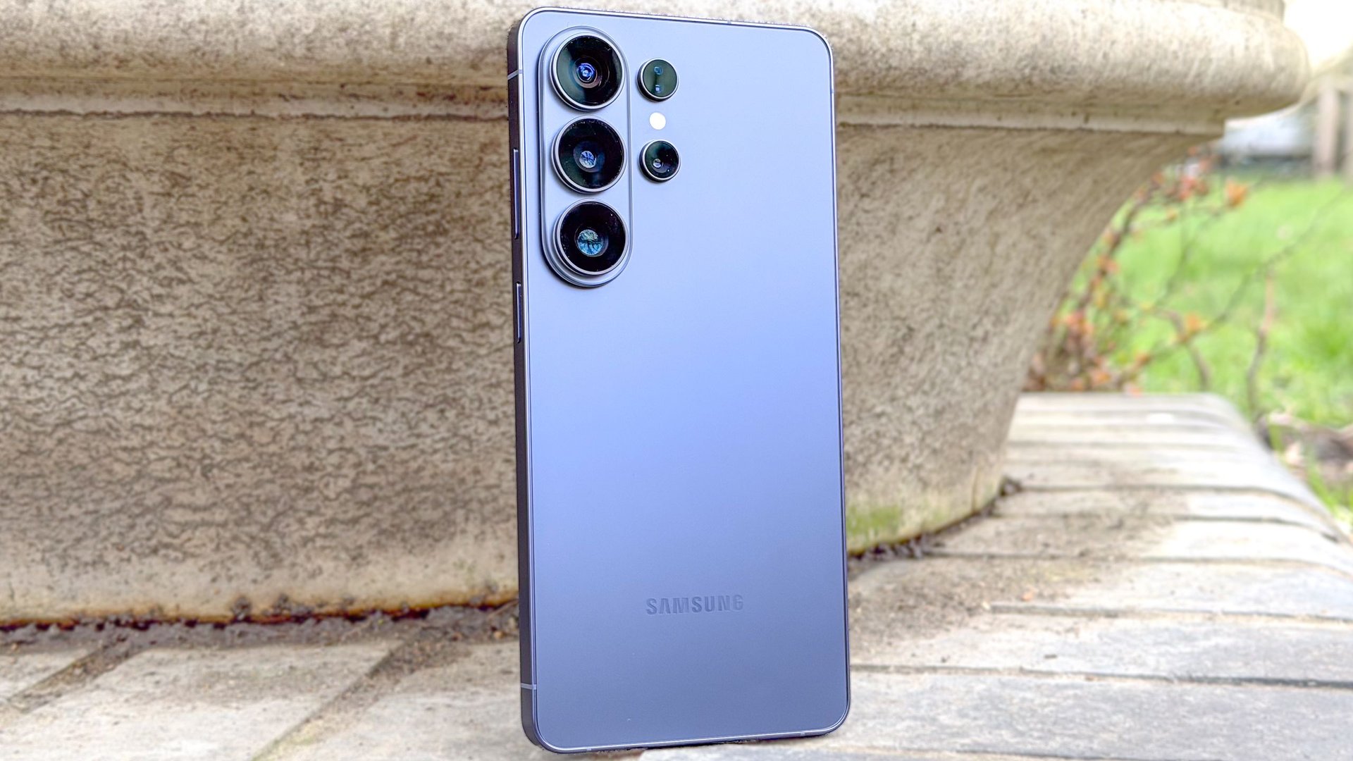 Blue Samsung smartphone standing upright on a stone surface showing a quad camera setup on the back