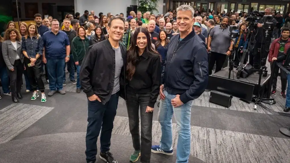 Three people pose at the front center of a large group photo smiling with attendees and camera crew in the background