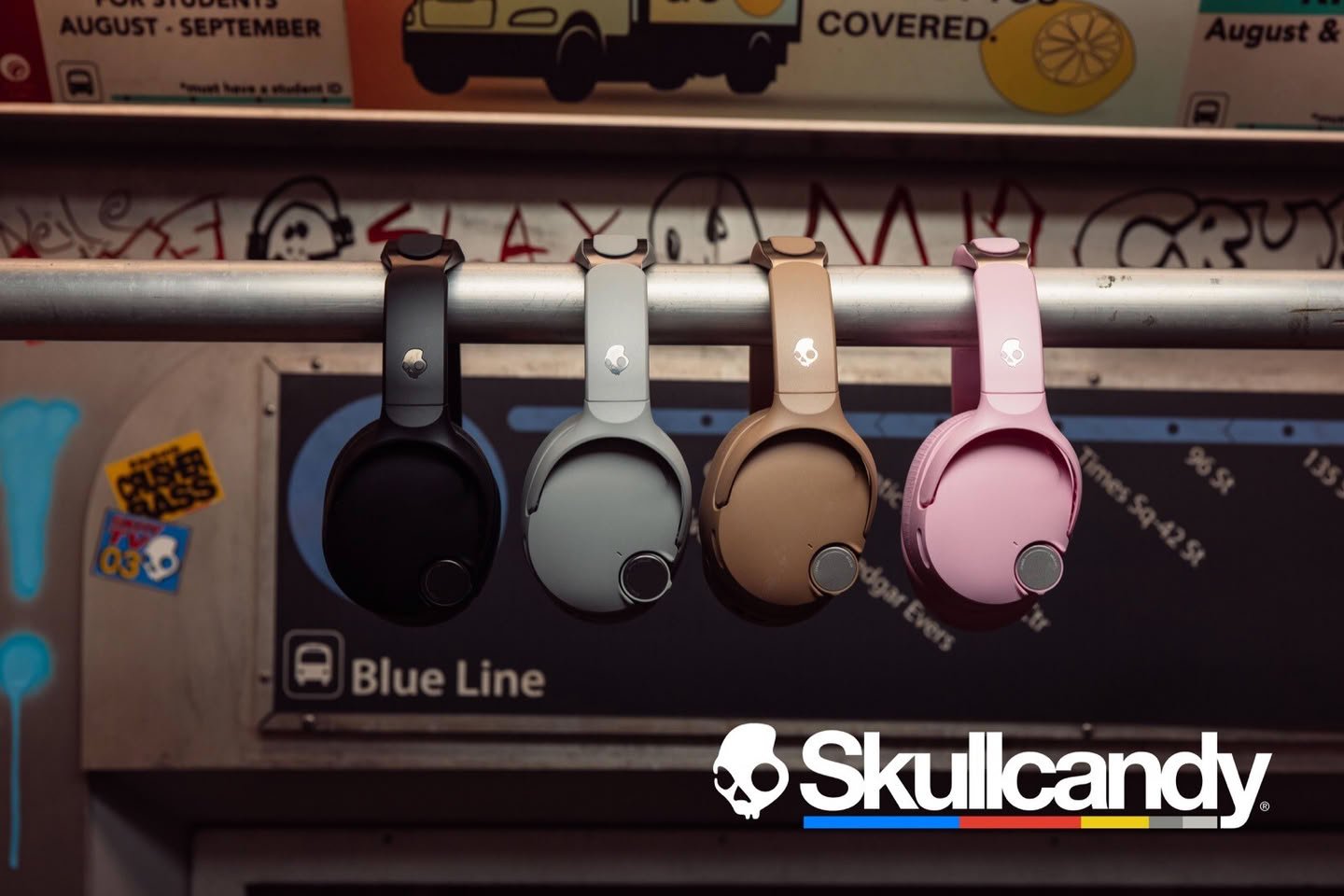 Skullcandy headphones in black gray tan and pink hanging on a rail in a subway car with a Blue Line sign in the background and the Skullcandy logo bottom right