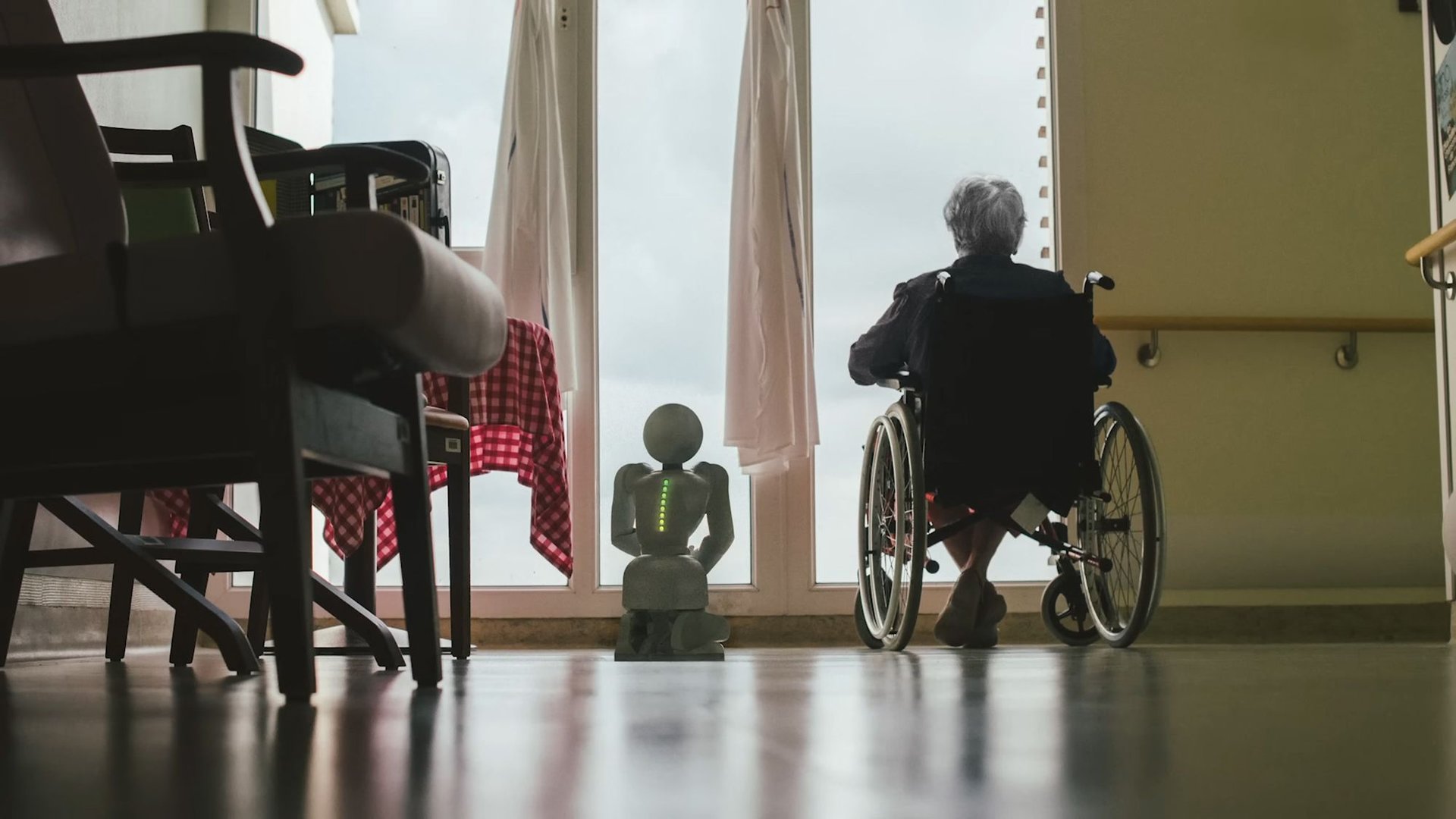 Person in a wheelchair looking toward a bright window with a small humanoid robot pedestal and green lights nearby in a care facility