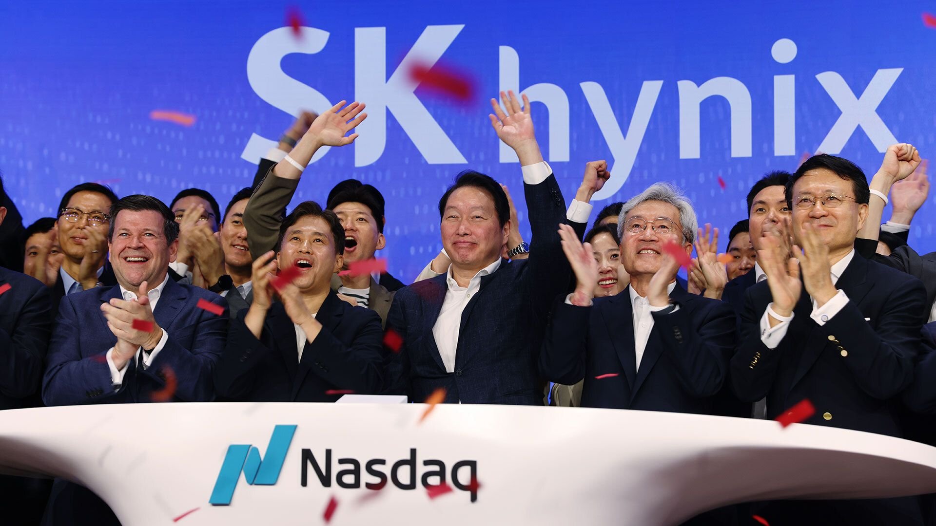 Group of diverse executives celebrating on stage at a Nasdaq event with confetti in the air and a blue backdrop displaying Nasdaq