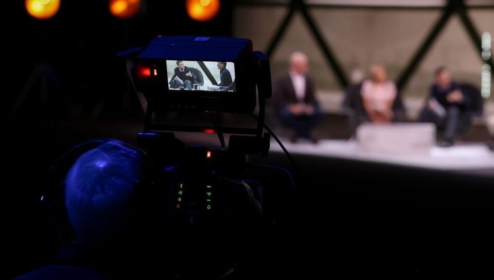 Video camera in foreground recording a panel discussion on screen monitor shows two speakers seated on stage with blurred audience in background